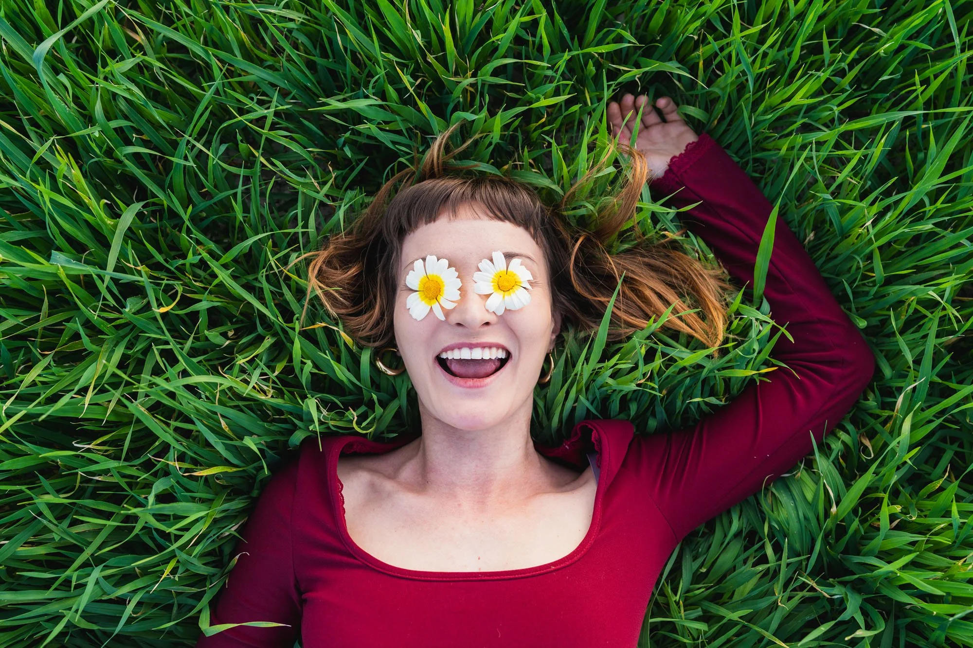 Mujer con cabello castaño claro con puntas rubias, usando camiseta roja, recostada en césped, con flores blancas y amarillas en los ojos, sonriendo ampliamente.