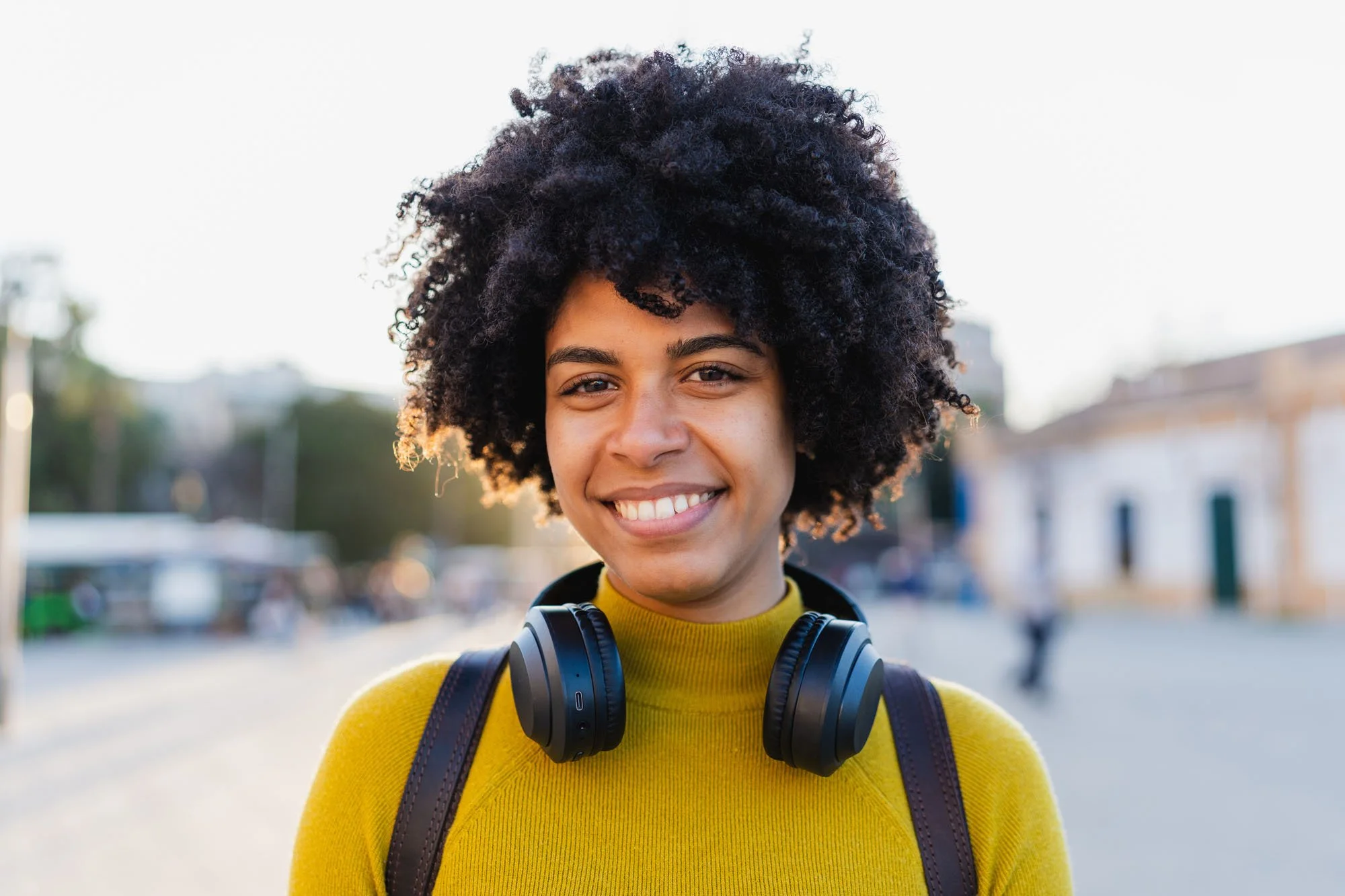 Mujer sonriente con cabello rizado en un entorno urbano, lleva audífonos, mochila y viste suéter amarillo.