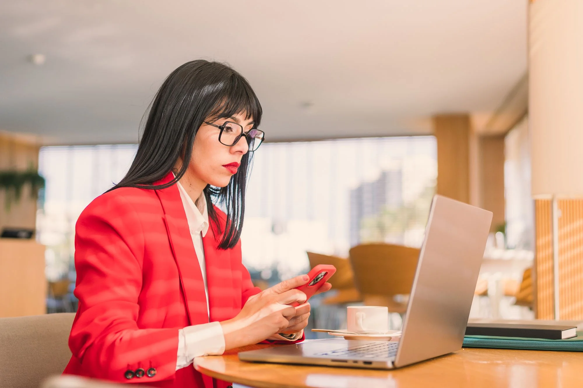 Mujer con gafas y cabello negro, vestida con saco rojo y blusa blanca, usando un teléfono móvil en una oficina moderna con ventanas grandes, computadora portátil y una taza en la mesa.