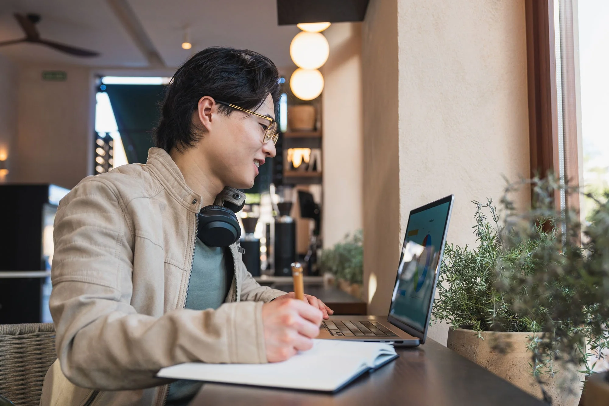 Joven con gafas y audífonos alrededor del cuello, escribiendo en un cuaderno mientras mira una computadora portátil sobre una mesa de madera junto a una ventana que permite la entrada de luz natural y plantas decorativas.