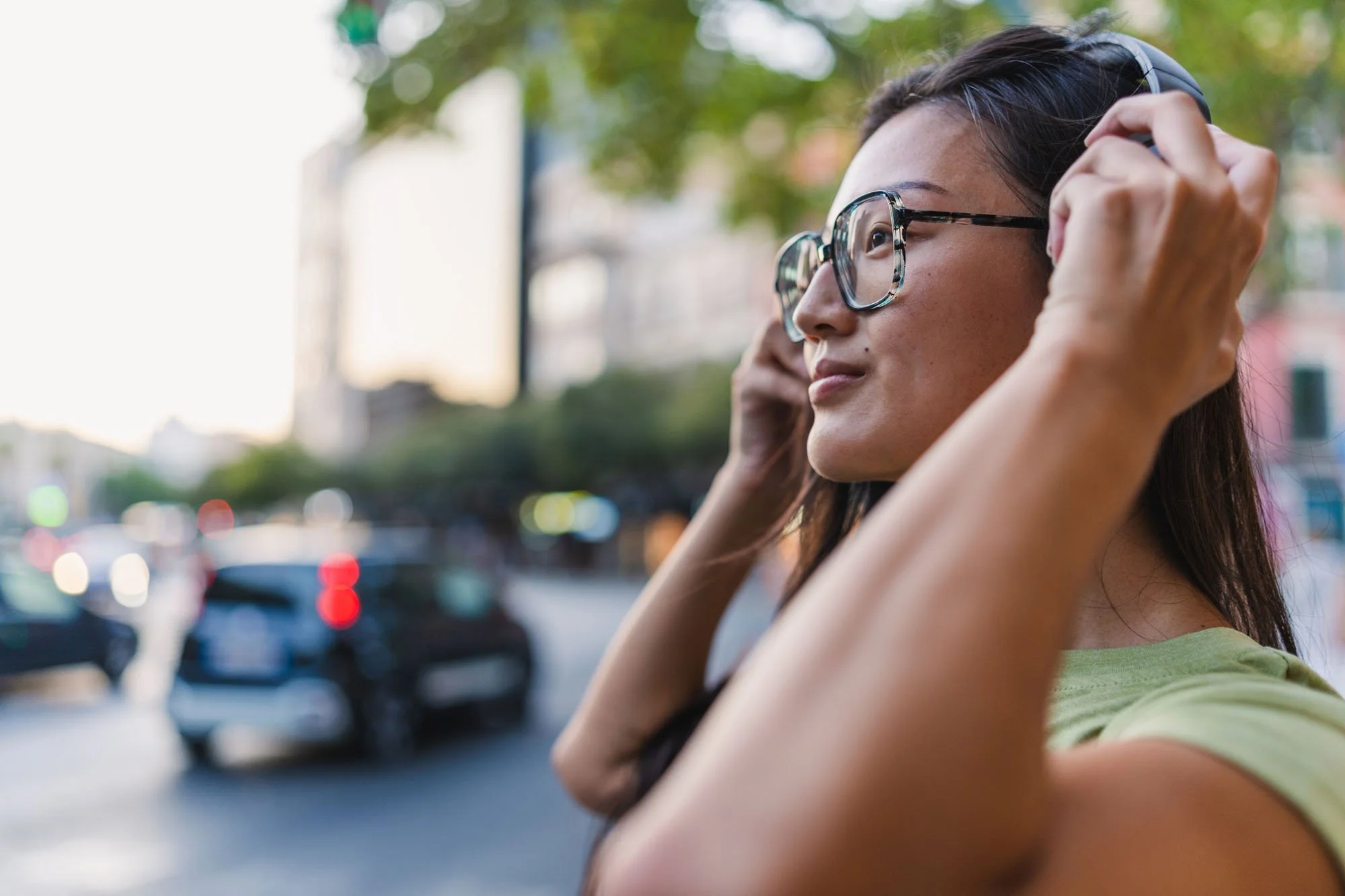 Mujer con gafas ajustándose los auriculares en un entorno urbano con árboles y vehículos de fondo.