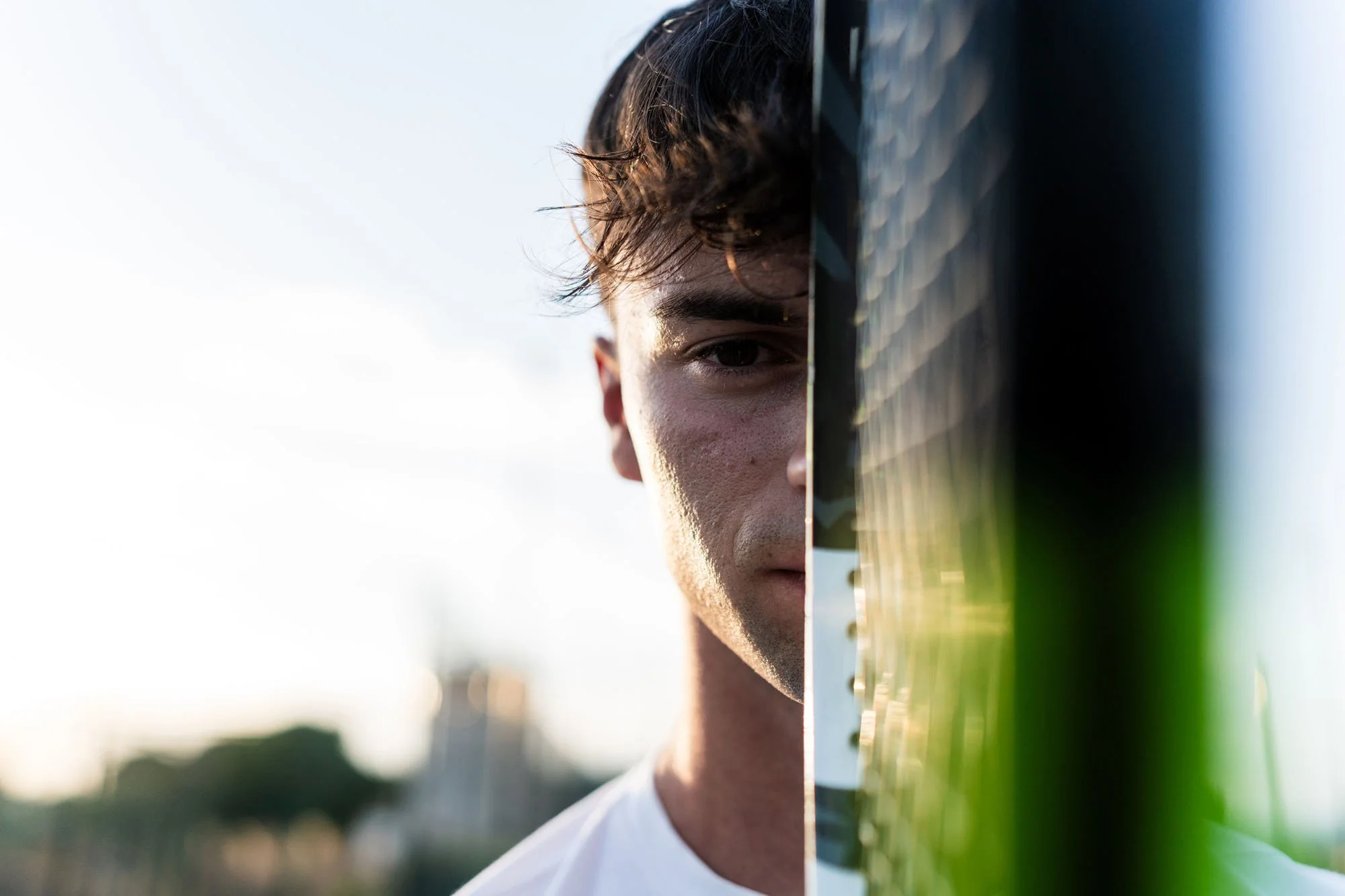 Retrato de un joven con rostro parcialmente visible, con cabello oscuro y ondulado, mirando hacia la cámara, con fondo neutro y superficie reflectante delante de su cara.