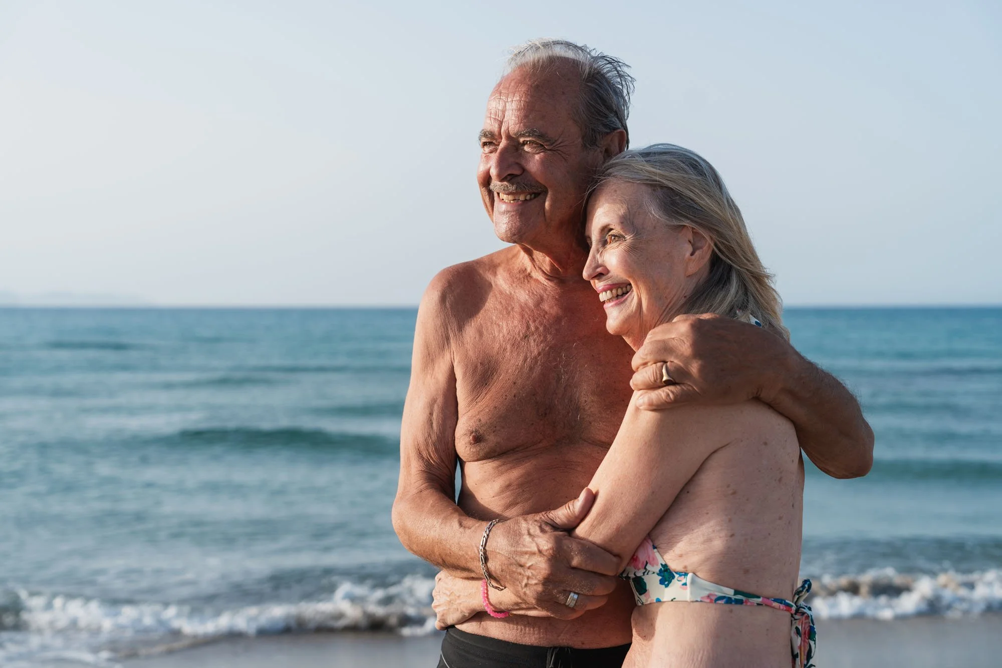 Pareja de ancianos abrazados en la playa, disfrutando del momento, con el mar y el cielo en el fondo.