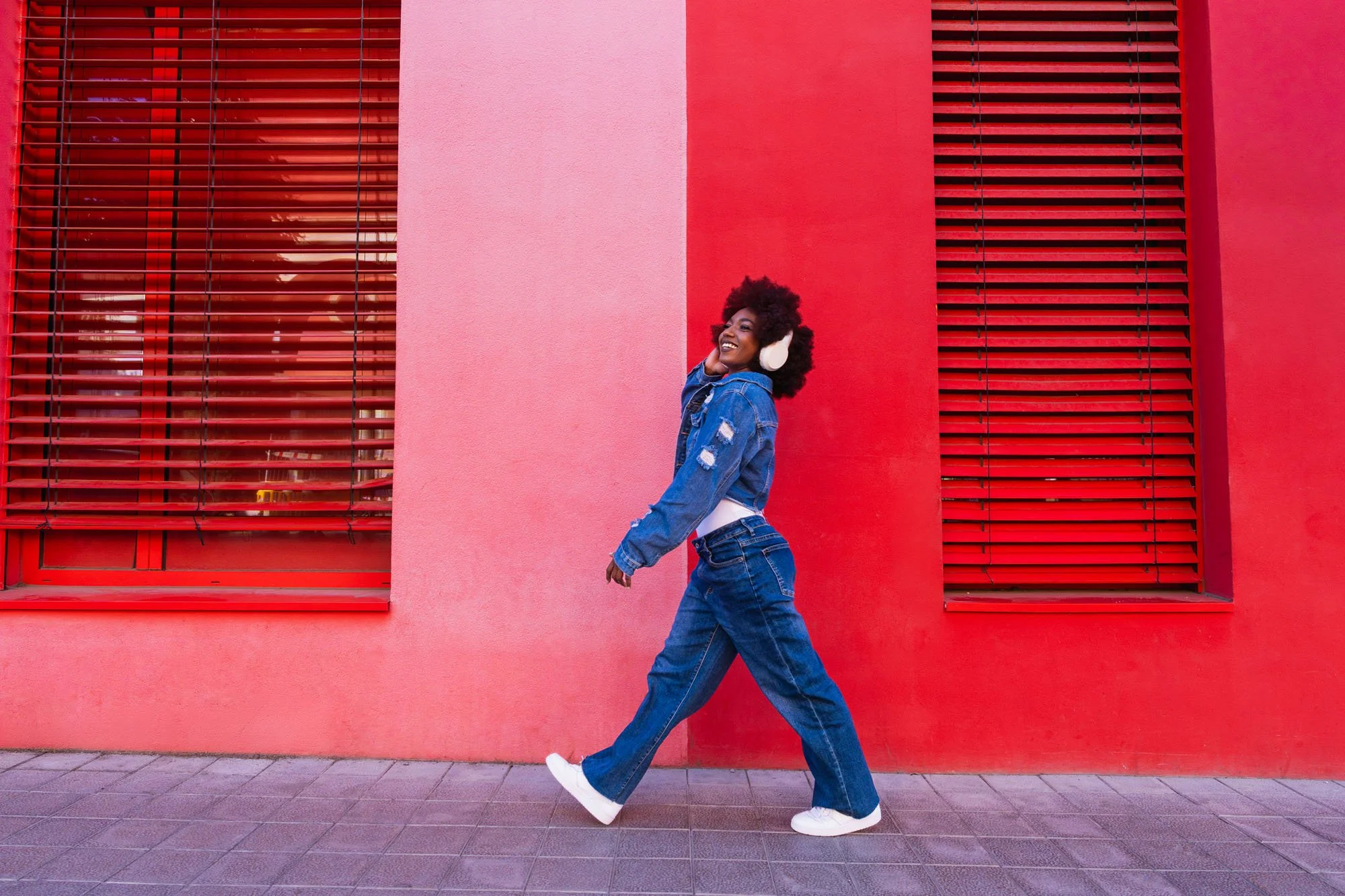 Mujer feliz con auriculares blancos, lleva abrigo de mezclilla con detalles rasgados y jeans, caminando frente a un muro rosa con ventanas rojas. 