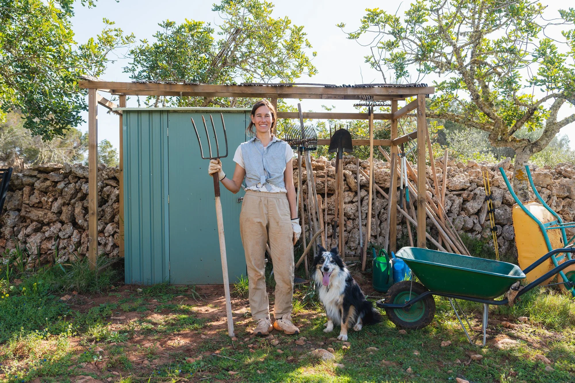 Una mujer con pantalones claros, una blusa de mezclilla y guantes sostiene una horca en un jardín rodeada de herramientas de jardinería y una perro pastor border collie.