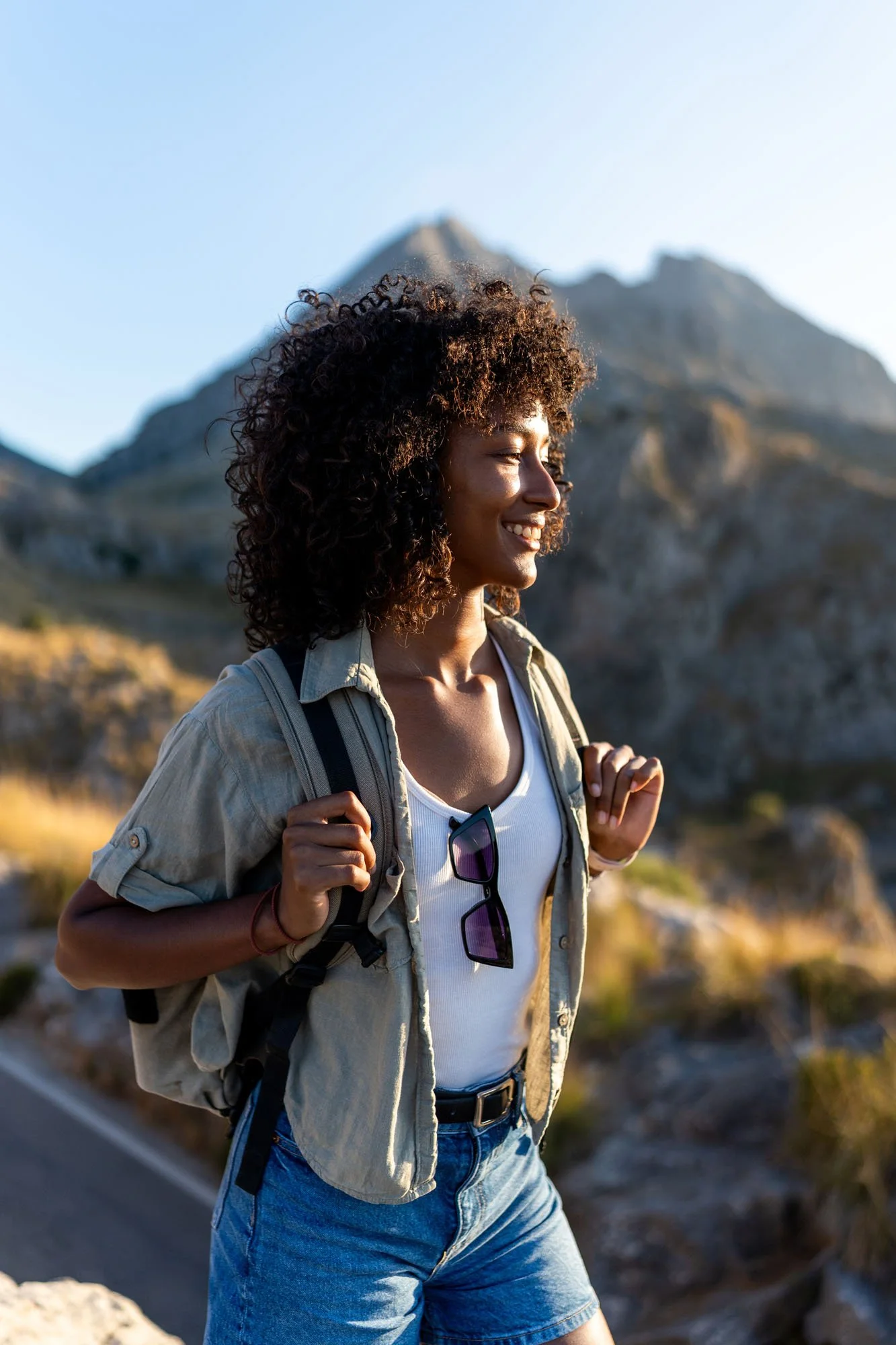 Joven mujer con cabello rizado, pantalones vaqueros y camiseta blanca, lleva una mochila y gafas de sol colgadas en la camiseta, caminando por un paisaje montañoso al atardecer.