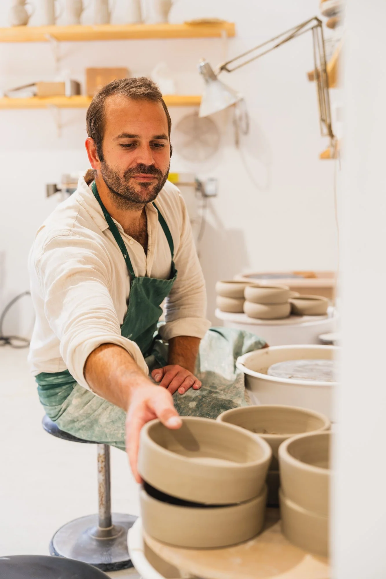 Un hombre joven, con barba y cabello corto, viste una camiseta blanca y un delantal verde, y está en un taller de cerámica colocando un recipiente de cerámica sin esmaltar. En el fondo hay estanterías con materiales y herramientas, y en la mesa hay v