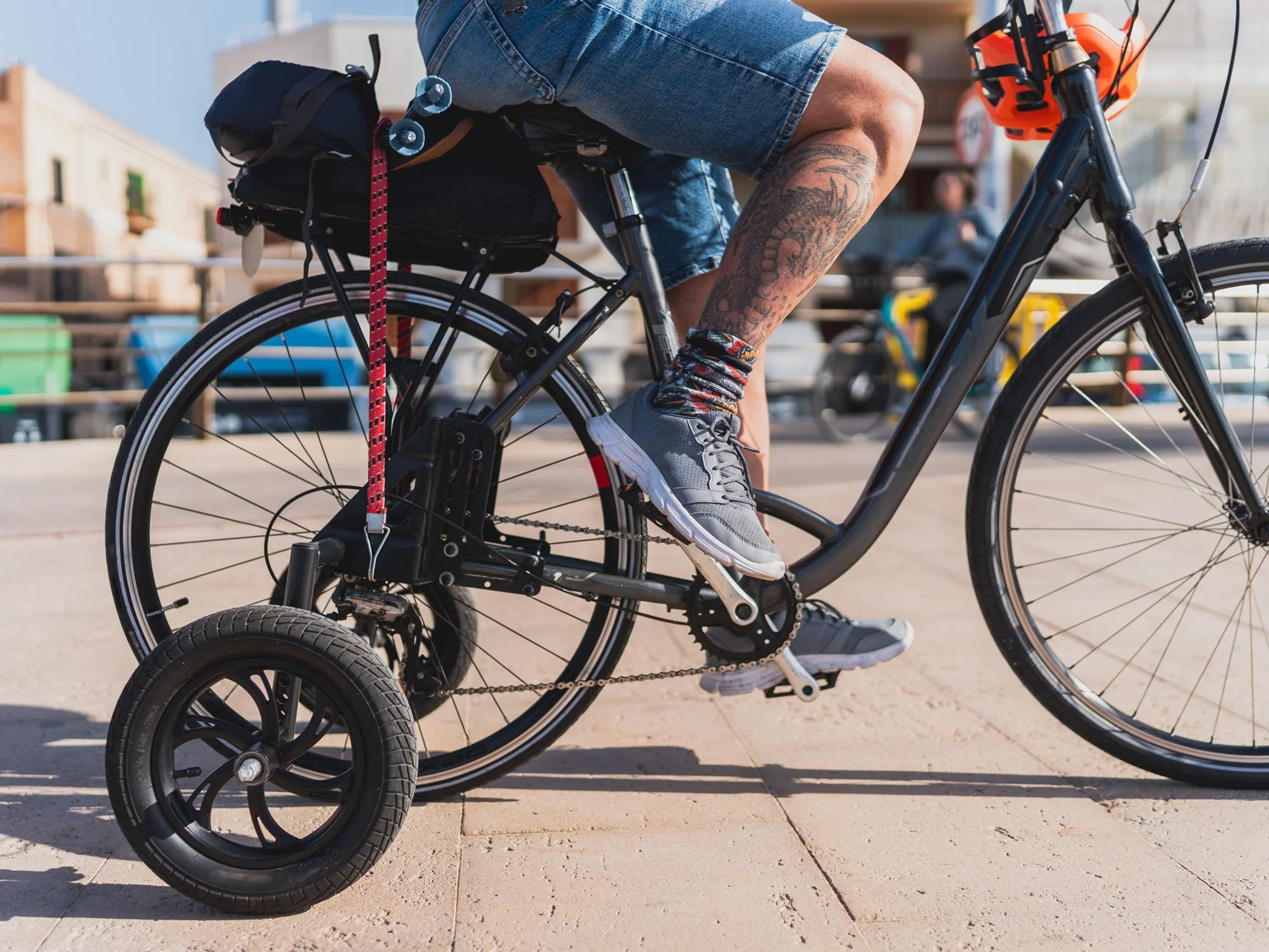 Persona montando una bicicleta eléctrica con llantas gordas en un espacio urbano, lleva pantalones cortos, zapatos deportivos y calcetas de colores. La bicicleta tiene accesorios y una mochila en el asiento trasero.