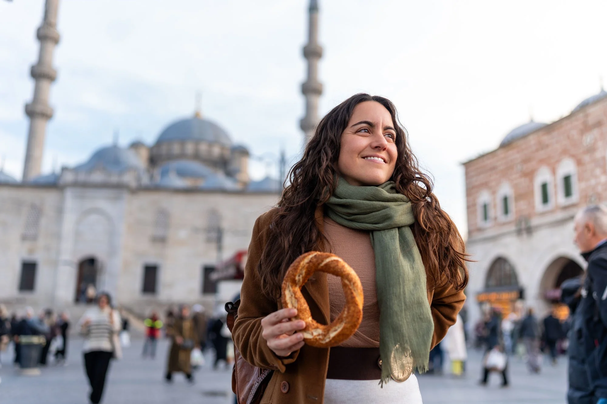Mujer con cabello rizado sosteniendo un pretzel grande en una plaza concurrida con arquitectura histórica y cúpulas en el fondo.