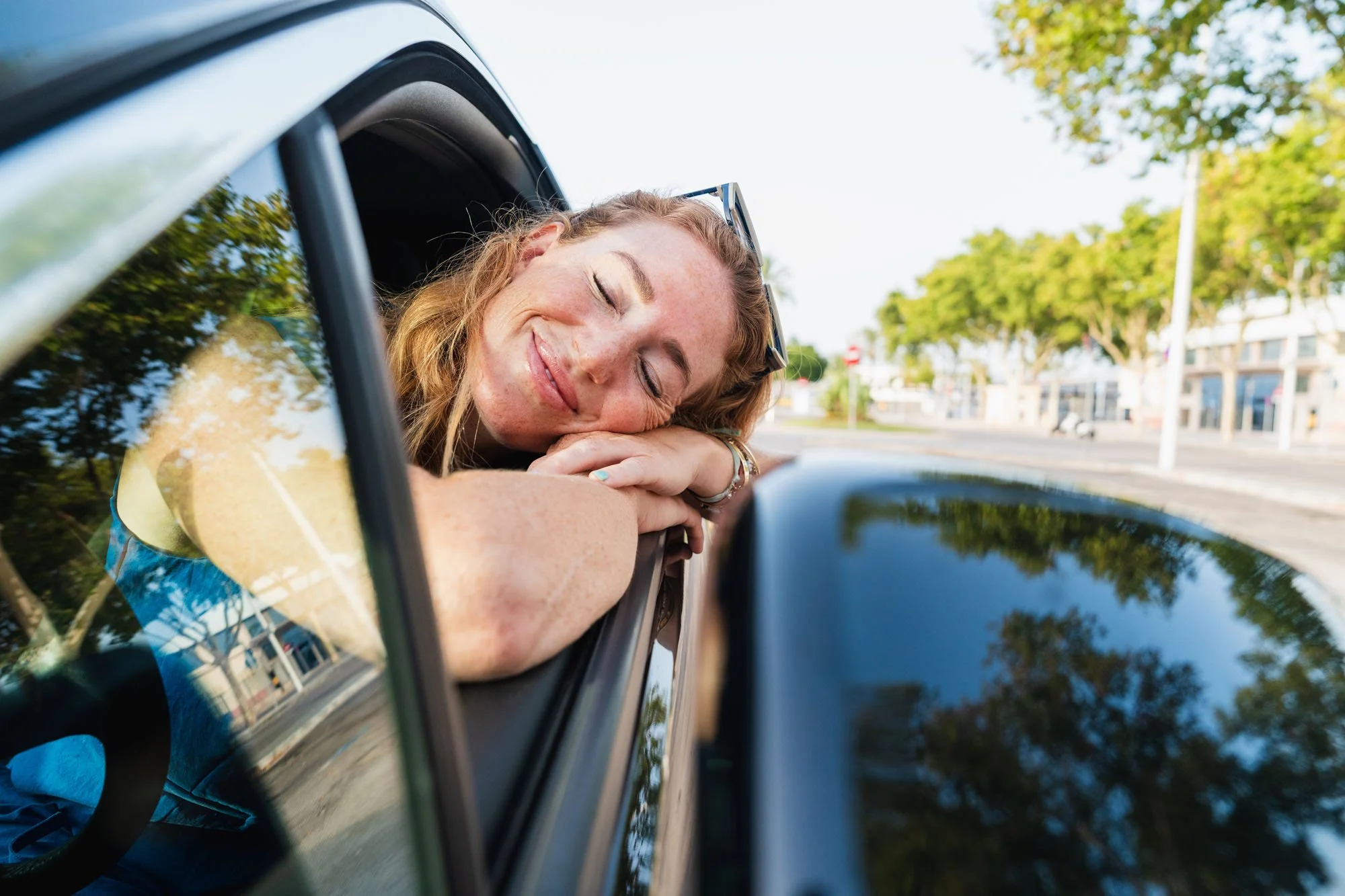 Mujer descansando con los ojos cerrados apoyada en la ventana de un automóvil en un día soleado en una calle con árboles y edificios