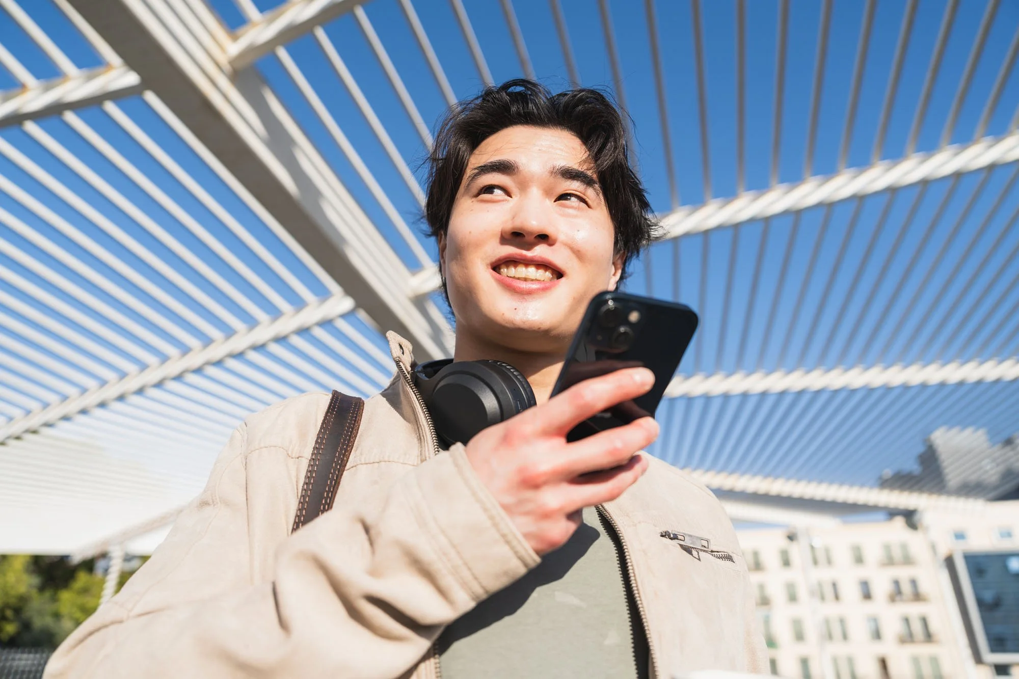 Joven hombre sonriente con auriculares alrededor del cuello usando un teléfono móvil en un espacio al aire libre con estructura metálica blanca y edificios en el fondo.