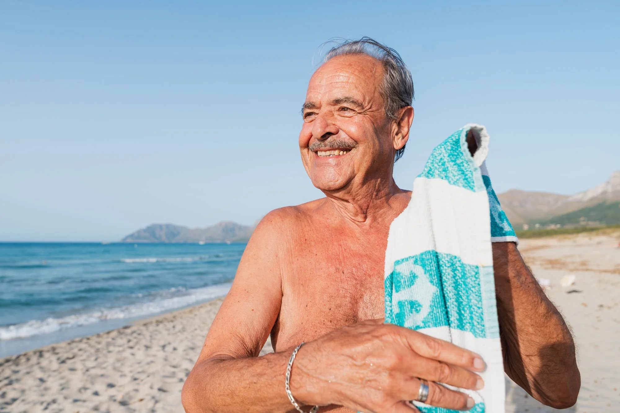 Hombre mayor sonriendo en la playa, sosteniendo una toalla, con el mar y montañas de fondo.