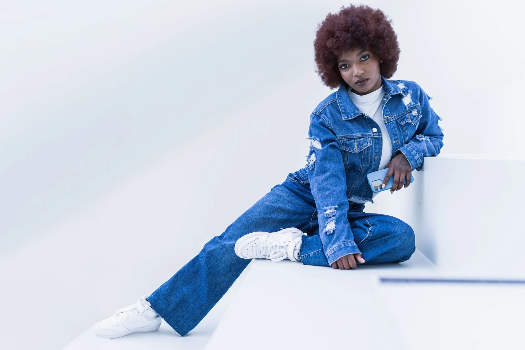 Mujer con cabello rizado pelirrojo, vestida con chaqueta y pantalón de mezclilla azules, sentada en una esquina blanca, sosteniendo un teléfono móvil, con zapatos blancos.