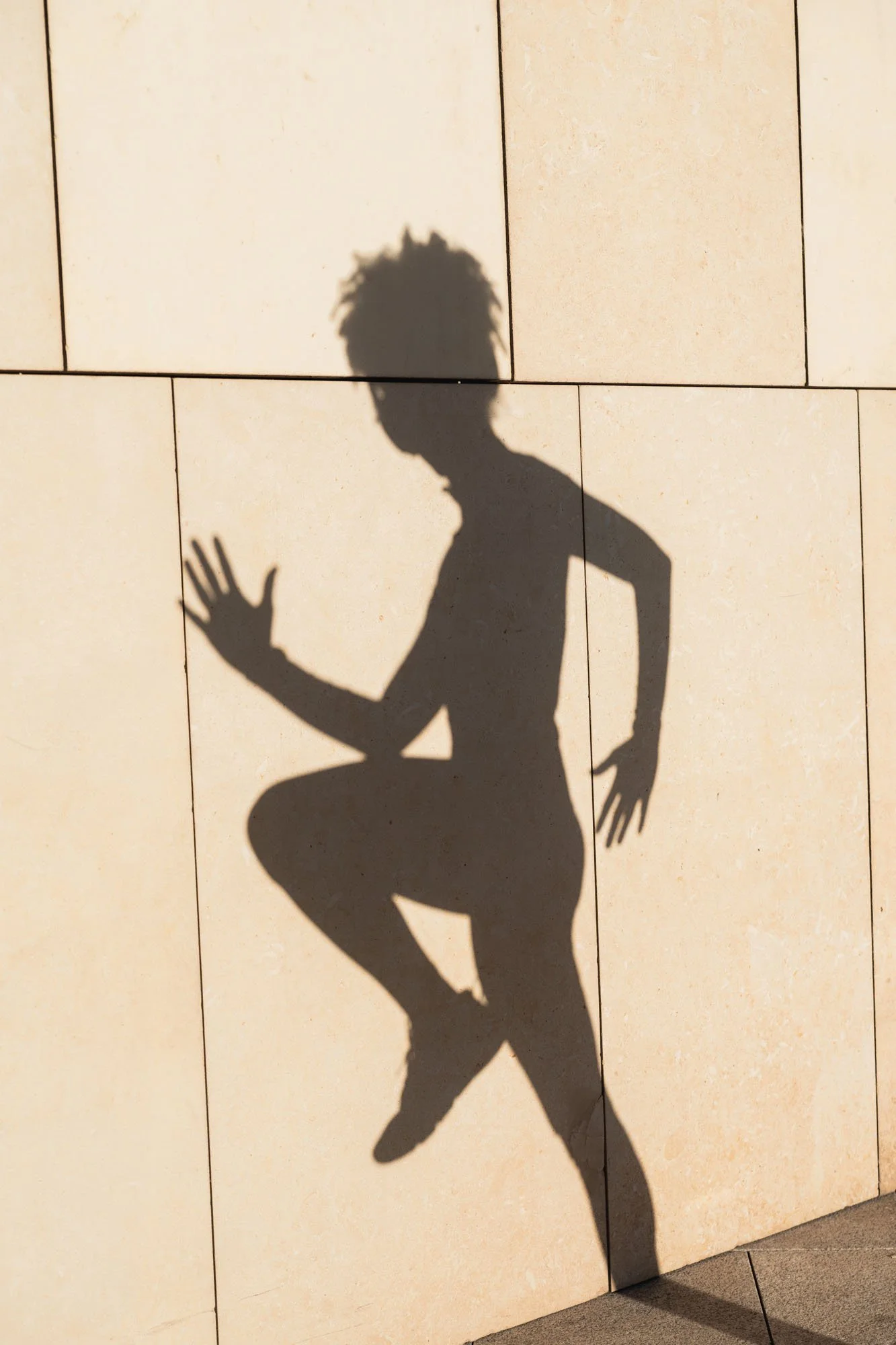 Sombra de una persona con peinado de dreadlocks haciendo un gesto de saludo con la mano en una pared de azulejos beige.