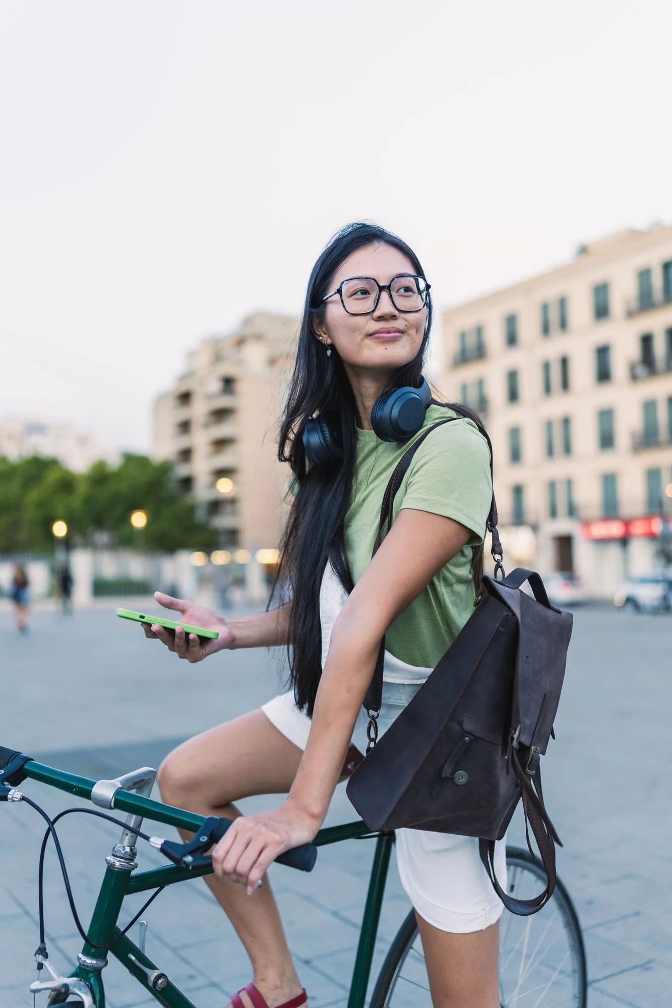 Joven mujer con gafas y auriculares en el cuello, montando bicicleta en ciudad, sosteniendo teléfono móvil, con edificios y árboles en el fondo.