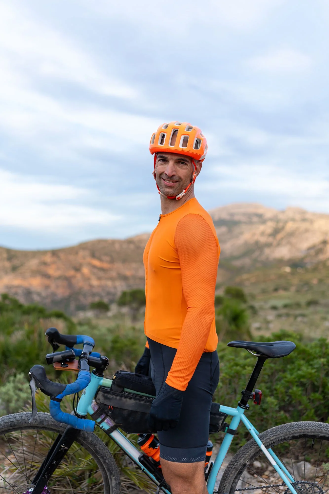 Hombre de pie con casco naranja y camiseta naranja, junto a bicicleta azul en paisaje montañoso con cielo nublado.