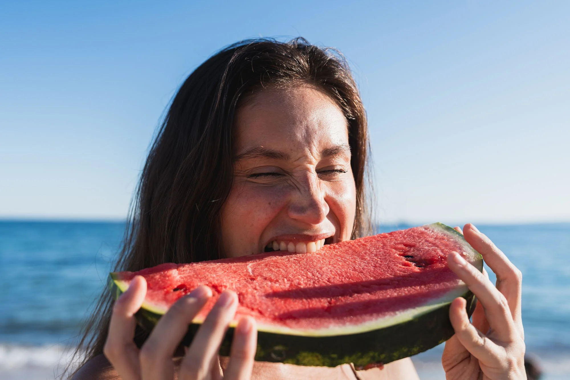 Mujer sonriendo con los ojos cerrados, sosteniendo una rebanada de sandía en la playa con mar y cielo despejado en el fondo.