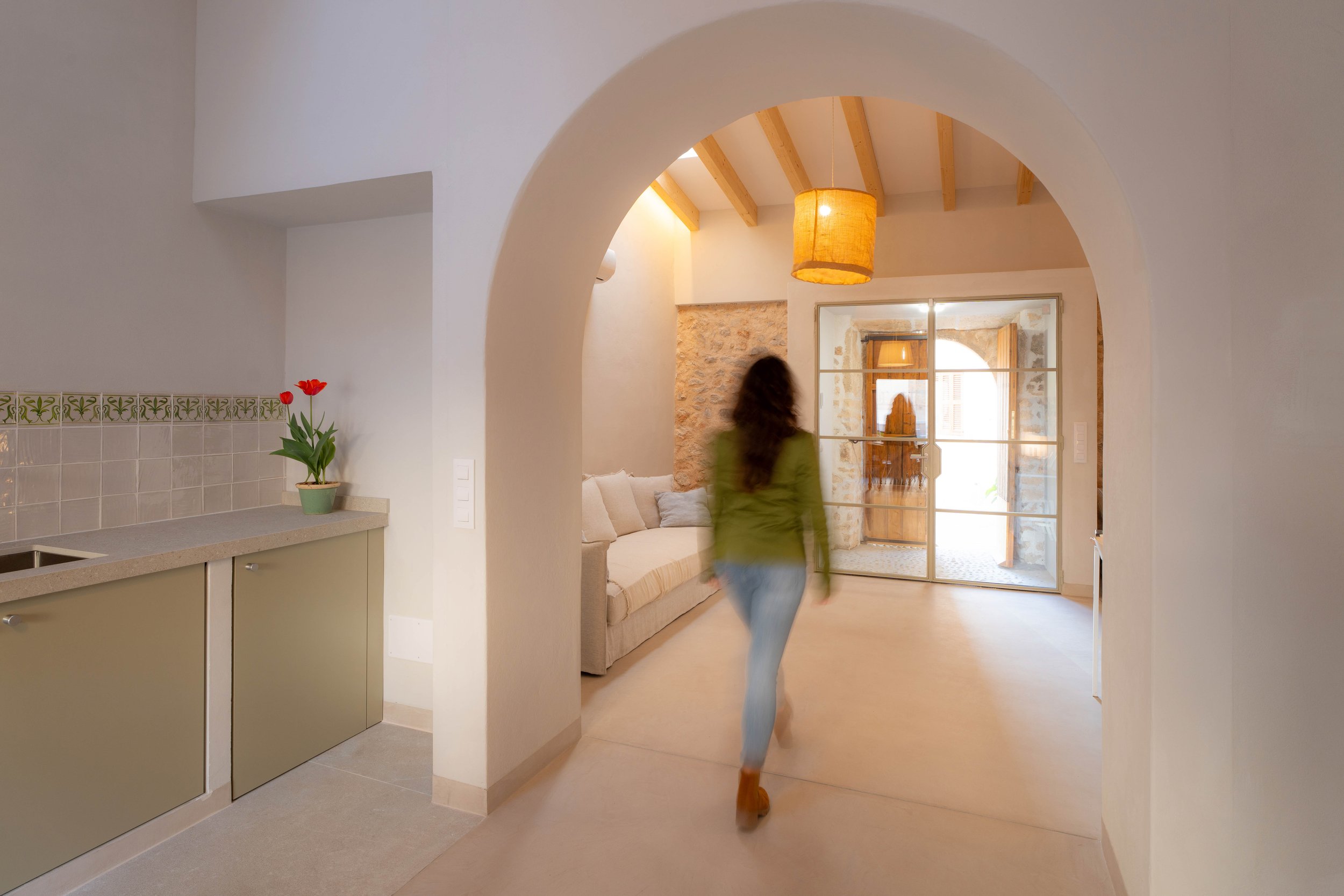 Interior de una casa con puerta de vidrio que da a un patio con paredes de piedra. Hay una sala con sofá y una mujer que camina con un bebé en brazos. En la cocina hay una planta de tulipanes rojos en una mesa.