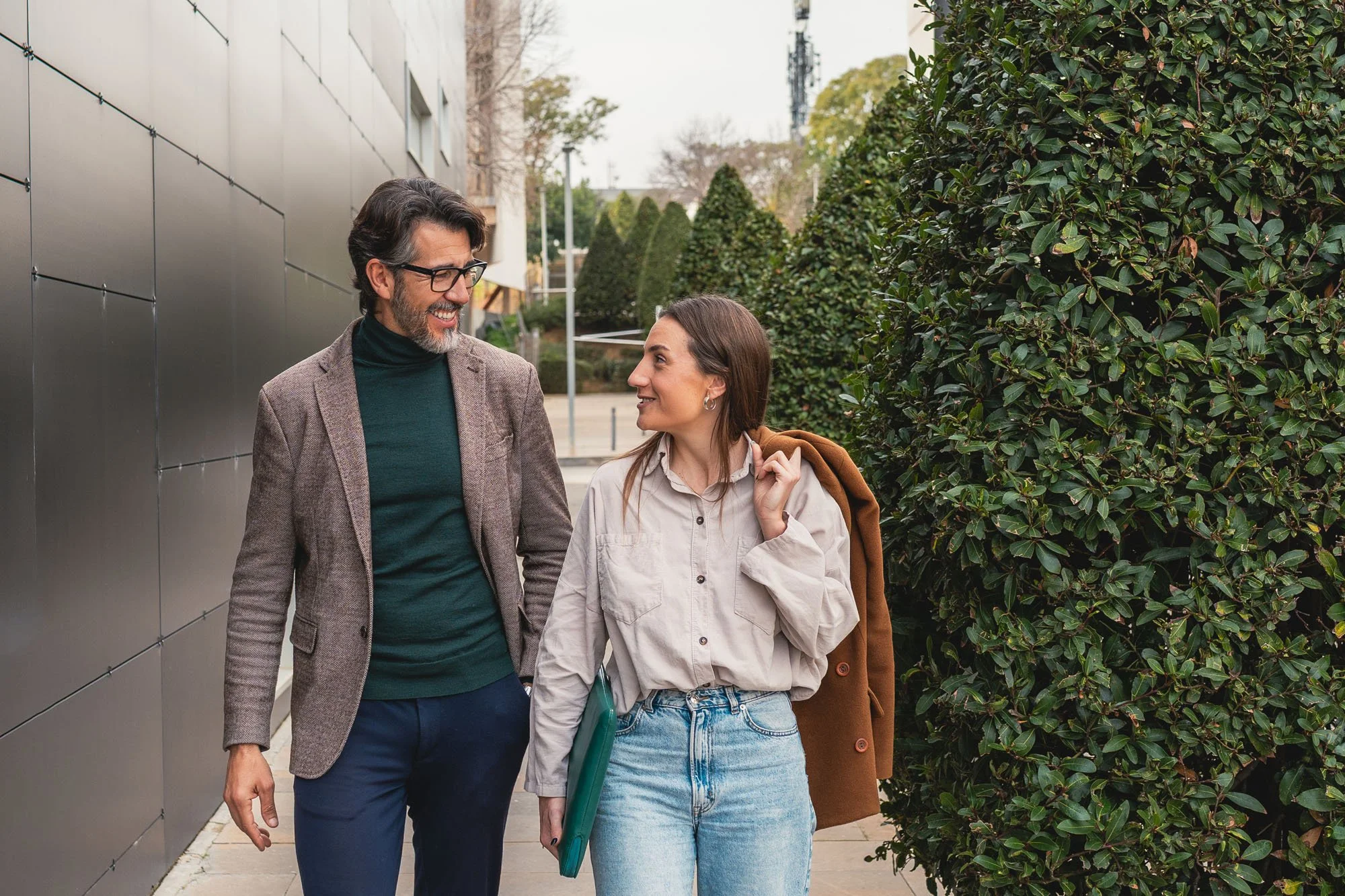 Dos personas conversando y caminando por un sendero rodeado de arbustos grandes en un día despejado.