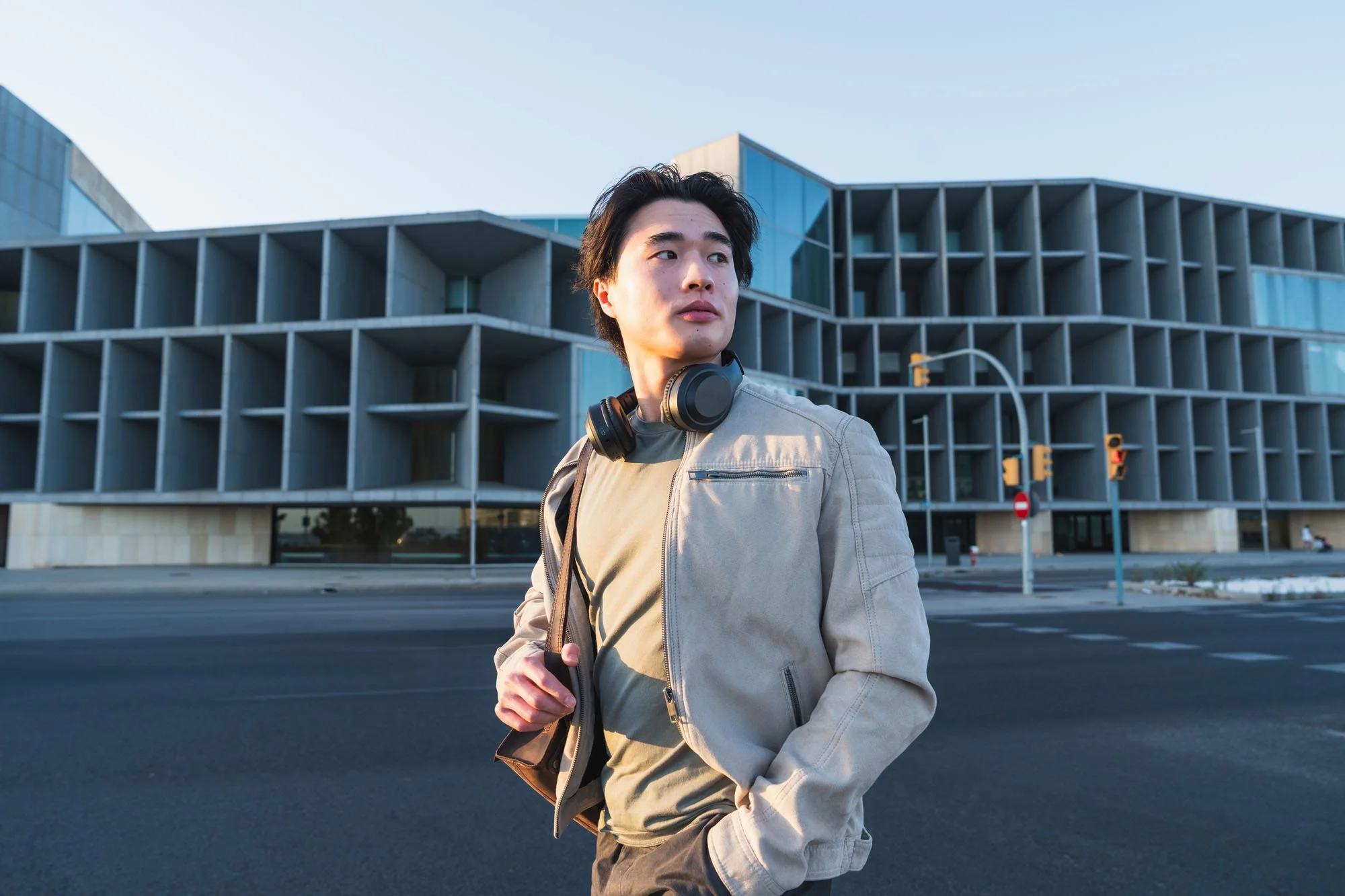 Joven con chaqueta y auriculares alrededor del cuello en ciudad moderna con edificios y semáforos.