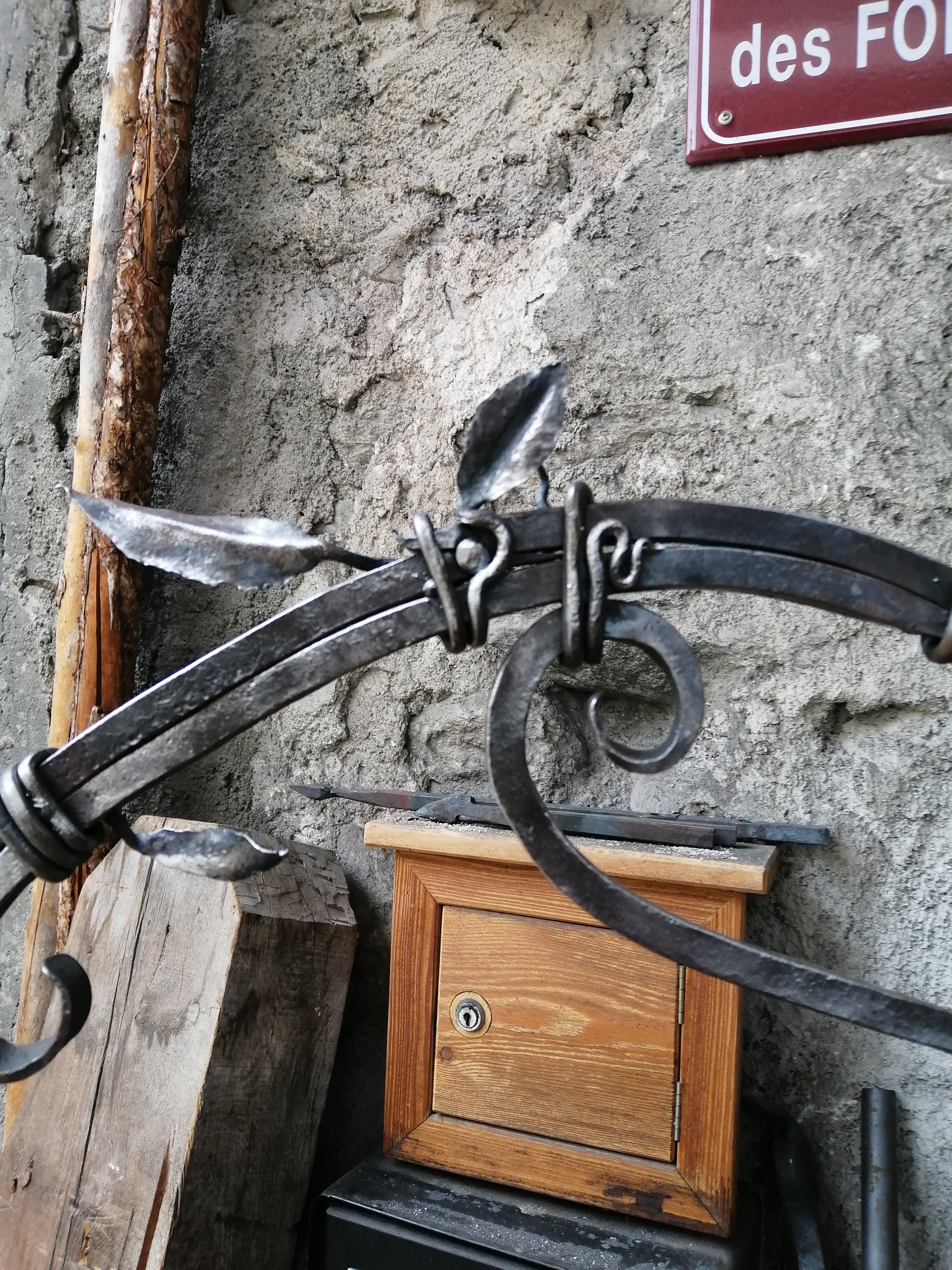 des décorations en métal forgé noir, un mur en pierre, une planche en bois à gauche, un panneau en métal rouge et blanc en haut à droite.