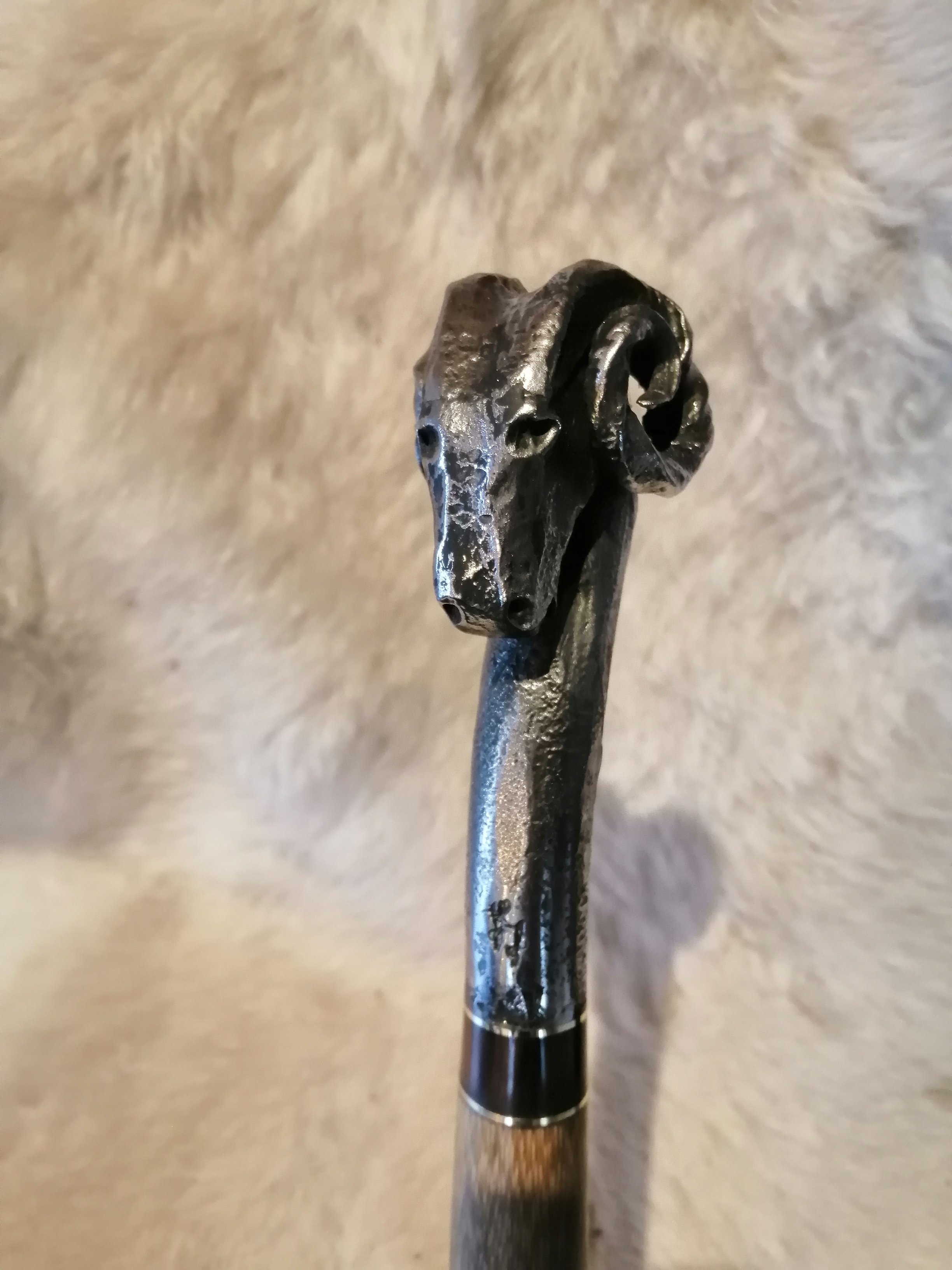 Un bâton de marche doté d'un morceau de bois en partie supérieur, avec une sculpture en forme de tête de bélier en fer forgé, posé sur un tapis en fourrure blanche.
