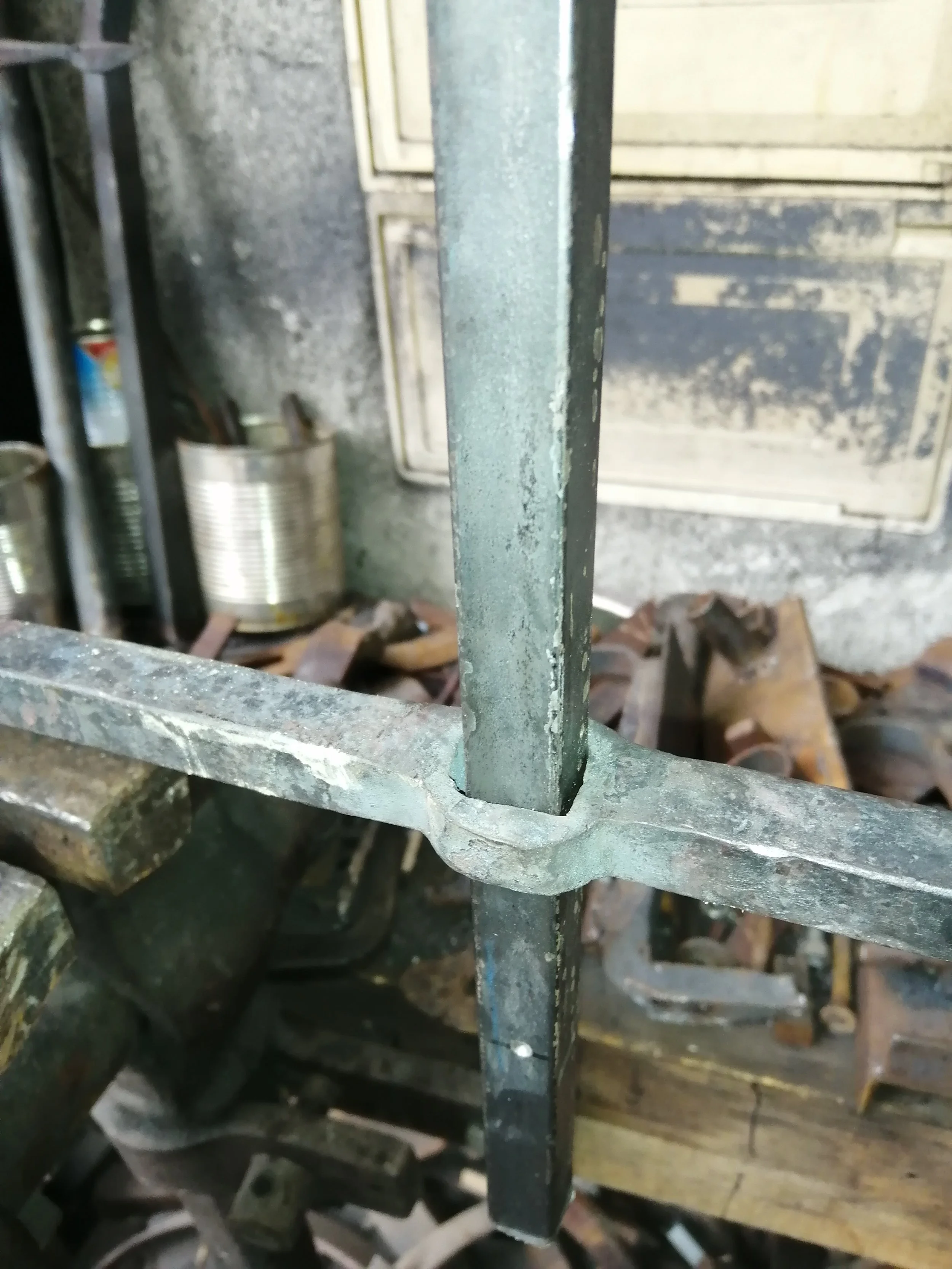 Une photo en gros plan d'une banque de fer forgé, probablement une barrière ou une grille, avec un objet noir posé dessus, dans un atelier ou un espace de bricolage, avec des outils et pièces métalliques en arrière-plan.