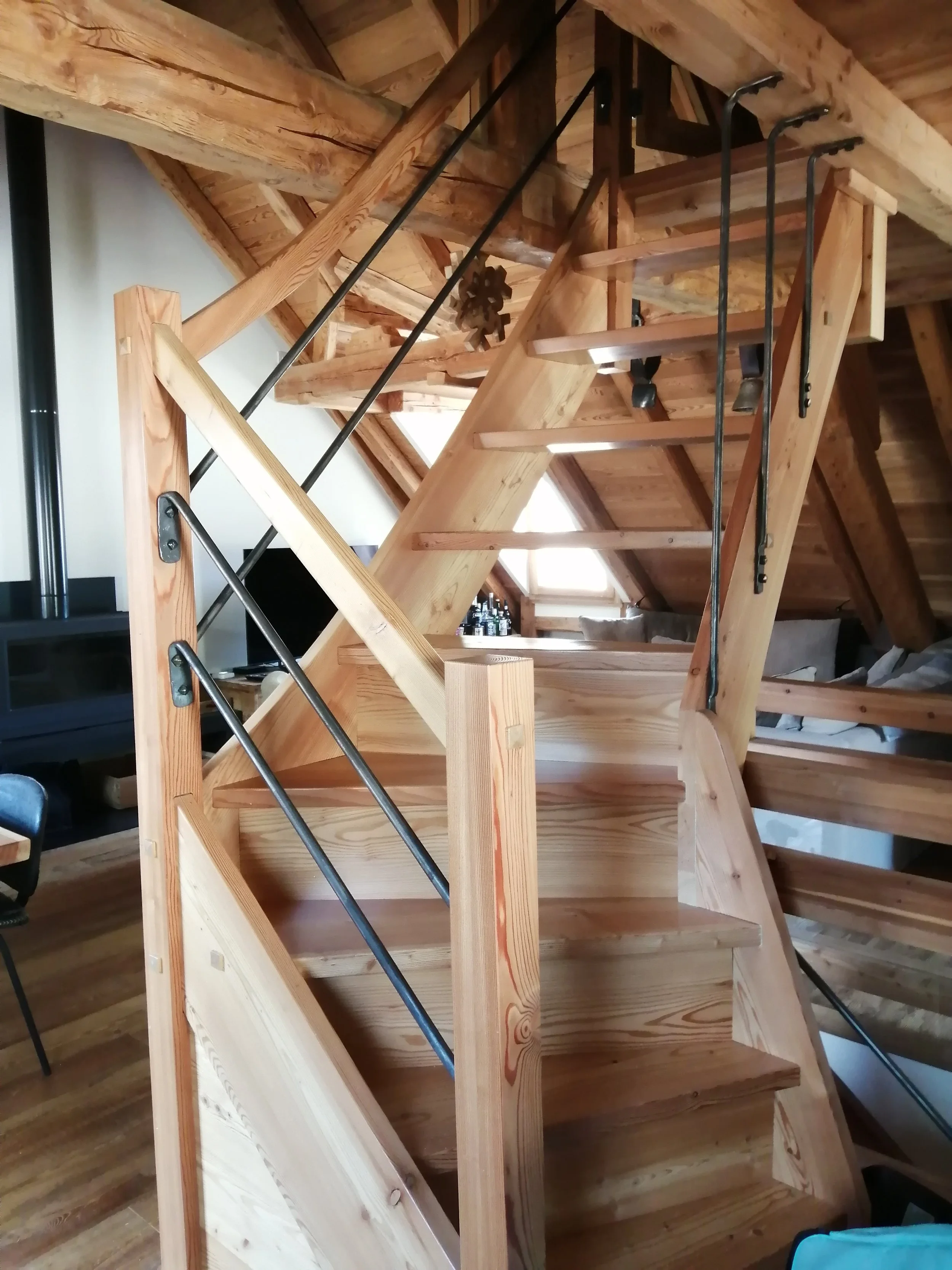 un escalier en bois dans une pièce avec un plafond en bois, éclairée par une lumière naturelle venant d'une fenêtre. les barreaux en fer forgé.