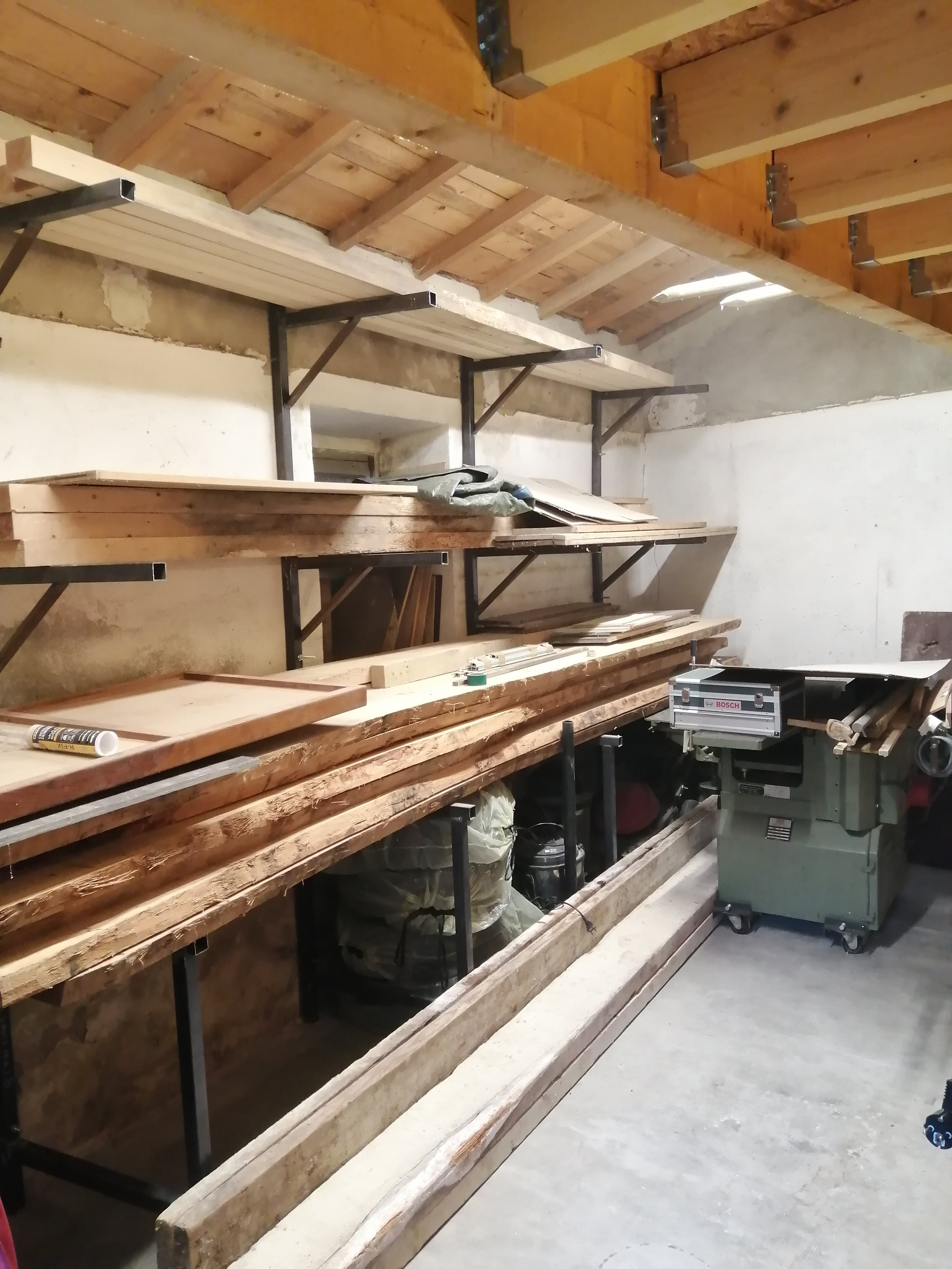 Un atelier de menuiserie avec des étagères en acier forgé, un espace de travail avec une scie à onglet et d'autres outils, disposition pour la fabrication ou la rénovation de meubles ou de structures en bois.