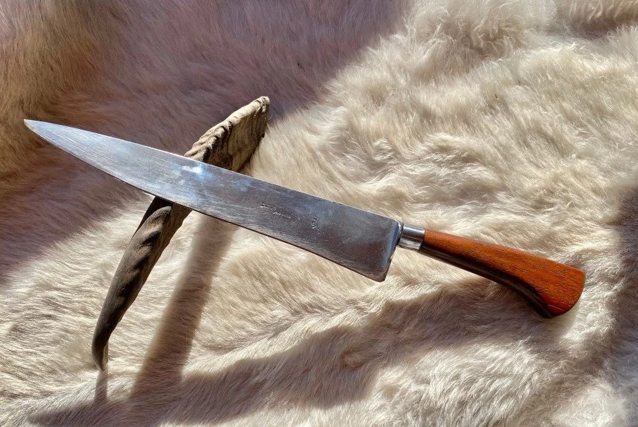 Un grand couteau de cuisine avec une lame en métal et un manche en bois, posé sur une surface en fourrure beige.