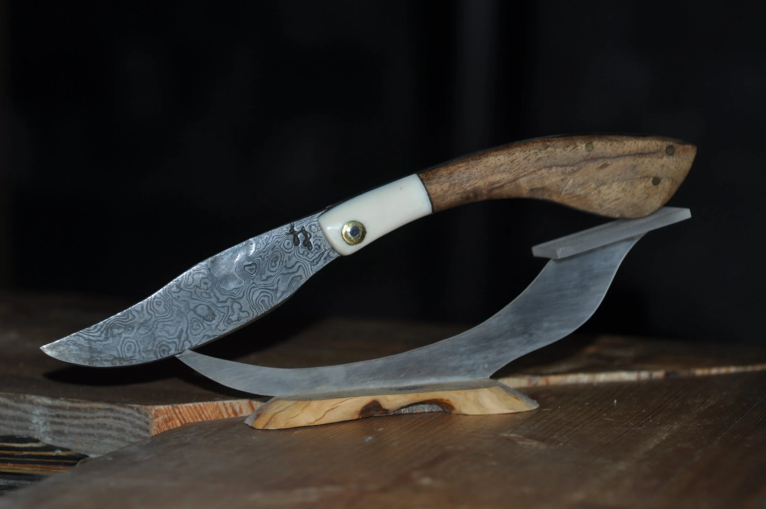 Couteau artisanal avec lame en damas et manche en bois et ivoire, posé sur une surface en bois avec un support en métal.