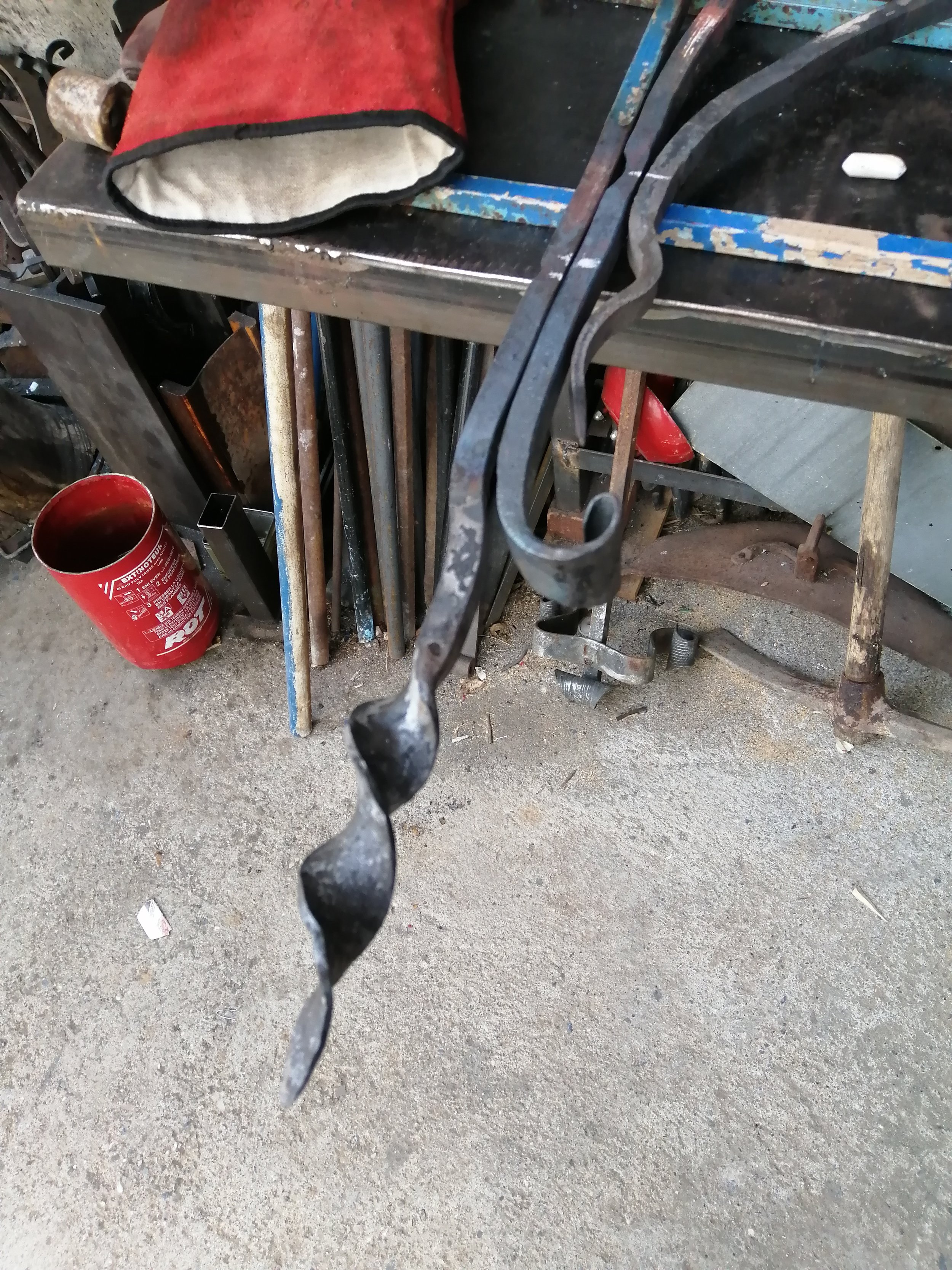 Un atelier avec des outils en métal, une escouette, un récipient rouge, des tubes métalliques et une table en métal travaillée. Le sol est en béton.