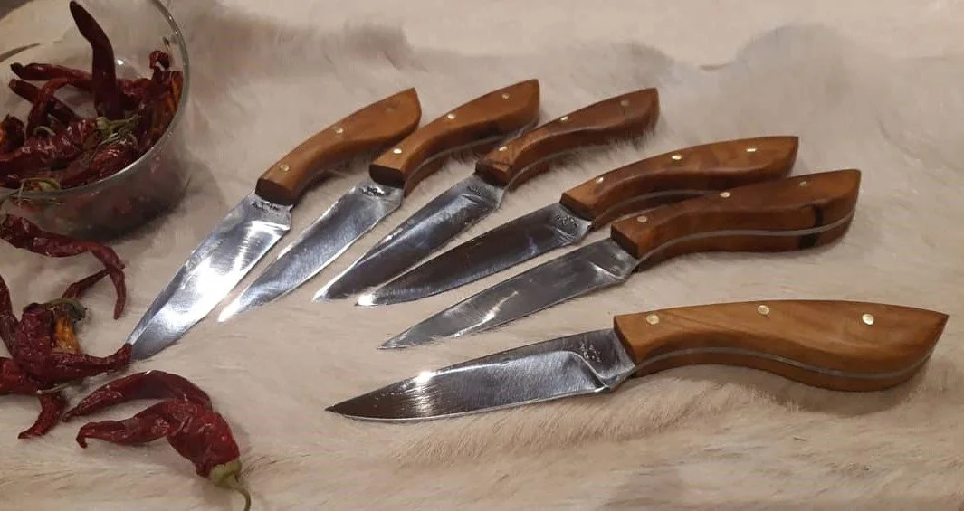 Six couteaux avec manches en bois et lames en acier carbone, disposés sur une surface blanche, à côté d'un bol contenant des piments rouges secs.