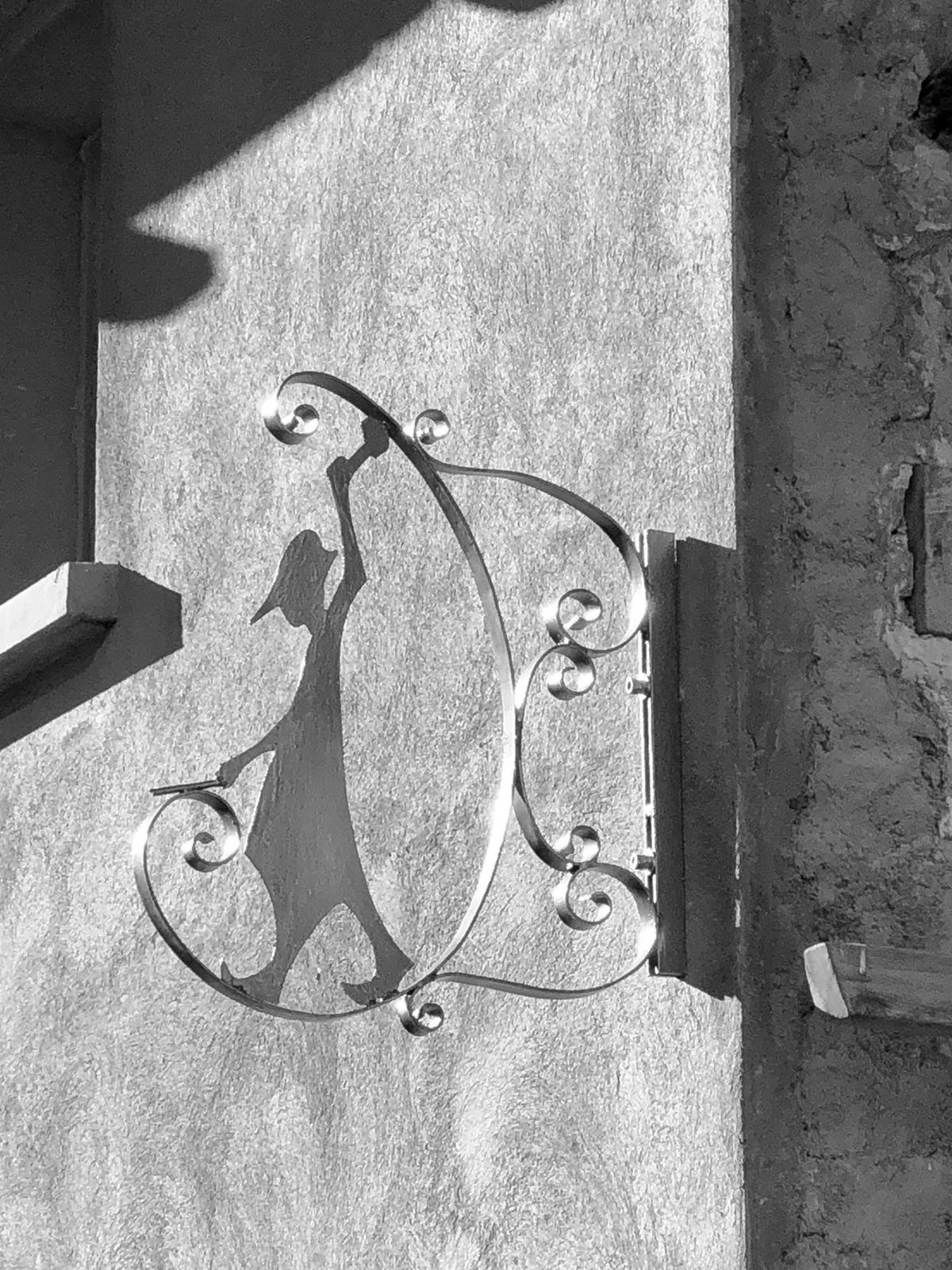 Une enseigne noire en fer forgé représentant la silhouette d'un forgeron mediéval portant un chapeau, fixée à un mur en pierre, avec un effet d'ombre projetée sur le mur.