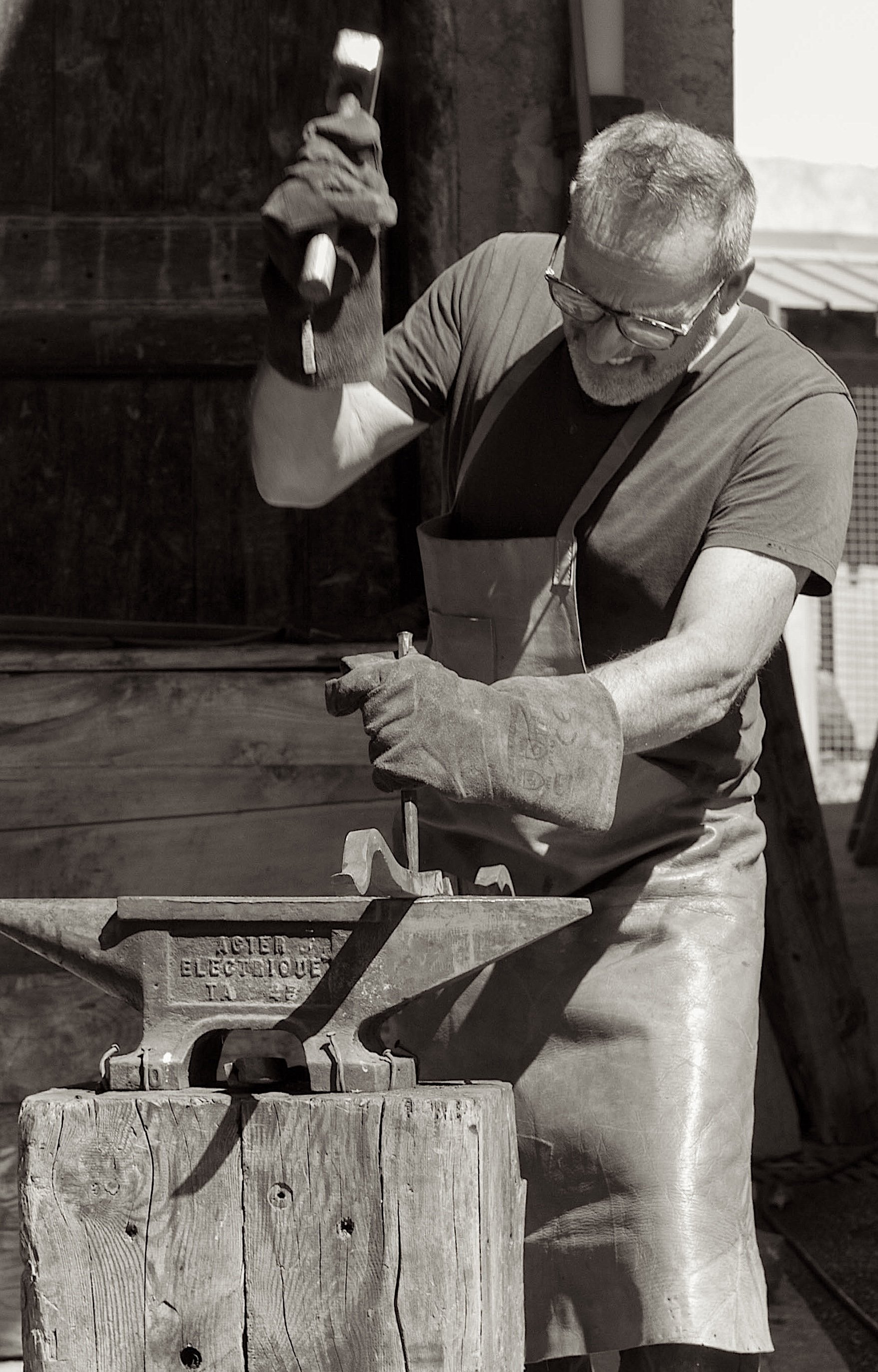 Un homme âgé avec des lunettes, portant un tablier, travaille avec un outil dans une forge. Il porte des gants et semble concentré sur son travail.