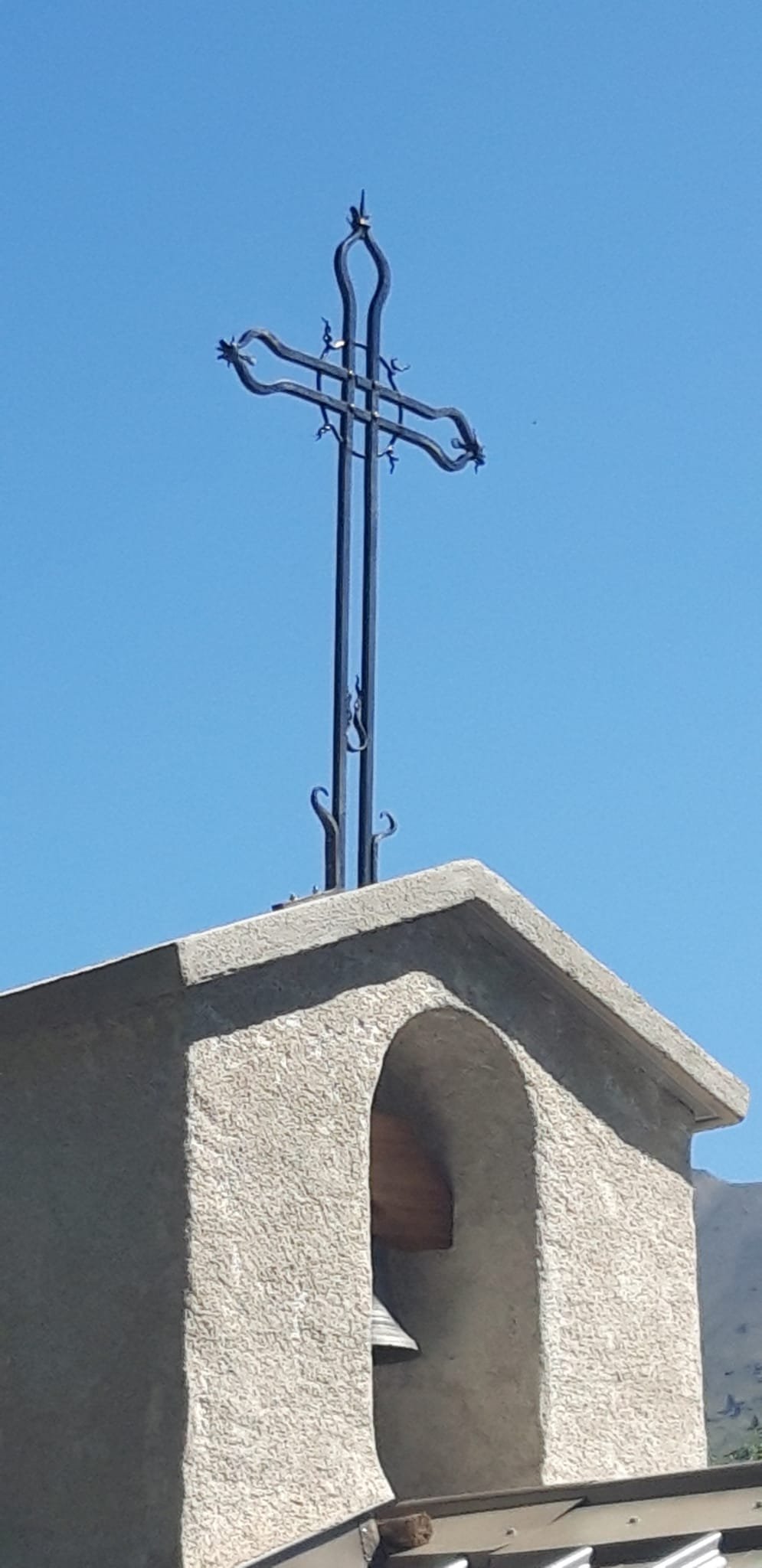 Une croix en métal fixée en haut d'une structure en pierre, contre un ciel bleu. La croix a une forme complexe et décorative.