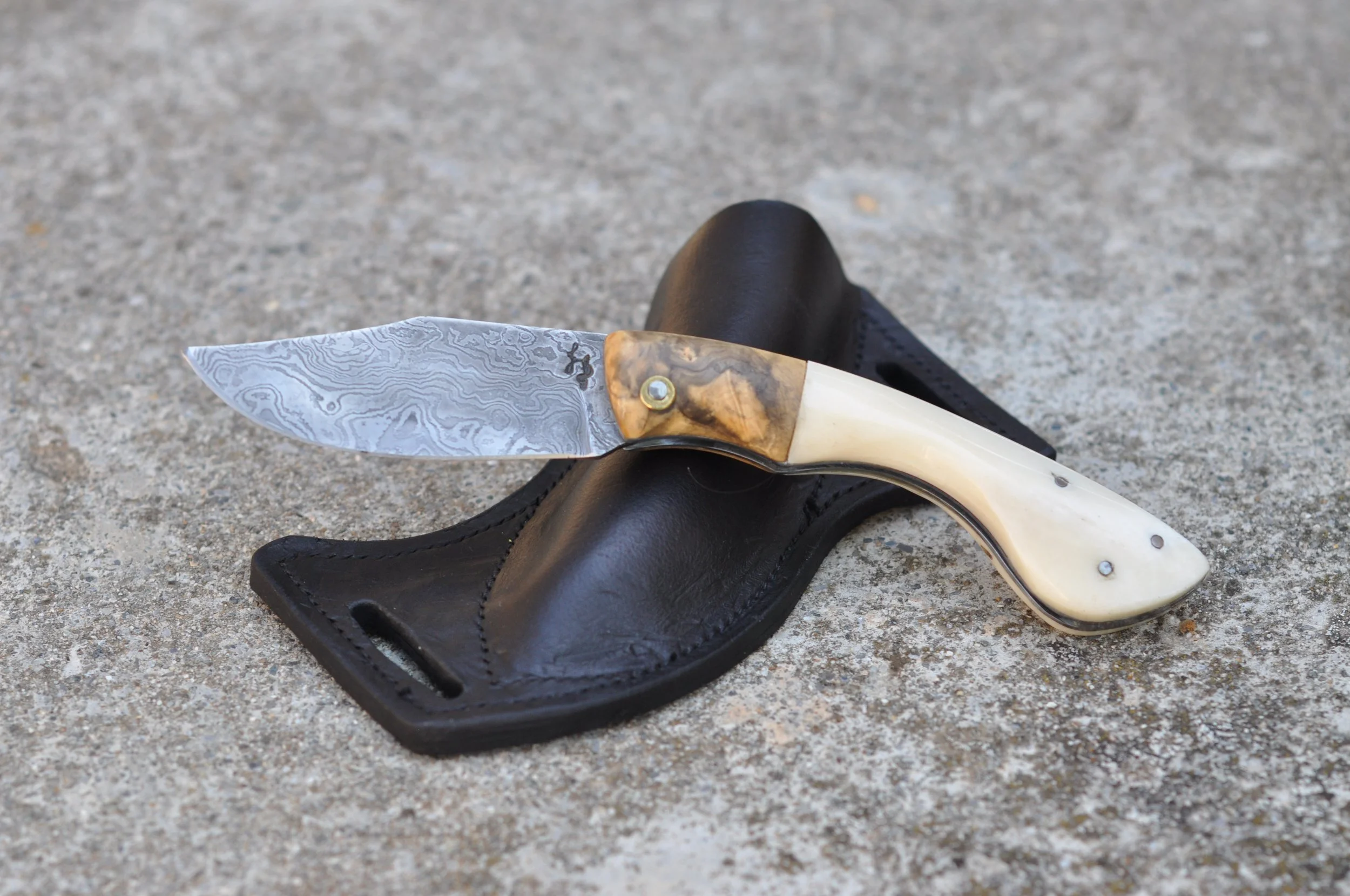 Un couteau à lame en acier aux motifs en damas, manche en ivoire ou imitation ivoire, posé sur un étui en cuir noir, sur un sol en béton ou pierre.