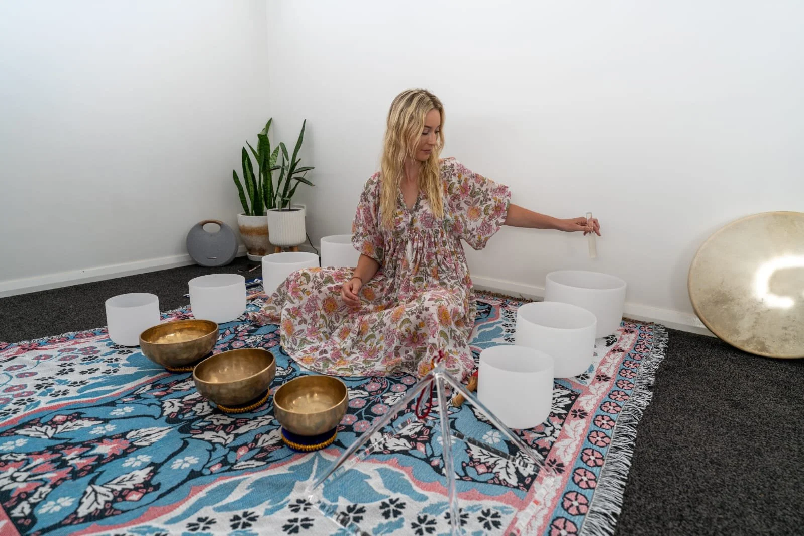A woman in a floral dress sitting cross-legged on a colorful rug, surrounded by singing bowls and a large cymbal, in a minimalistic room with white walls and potted plants.