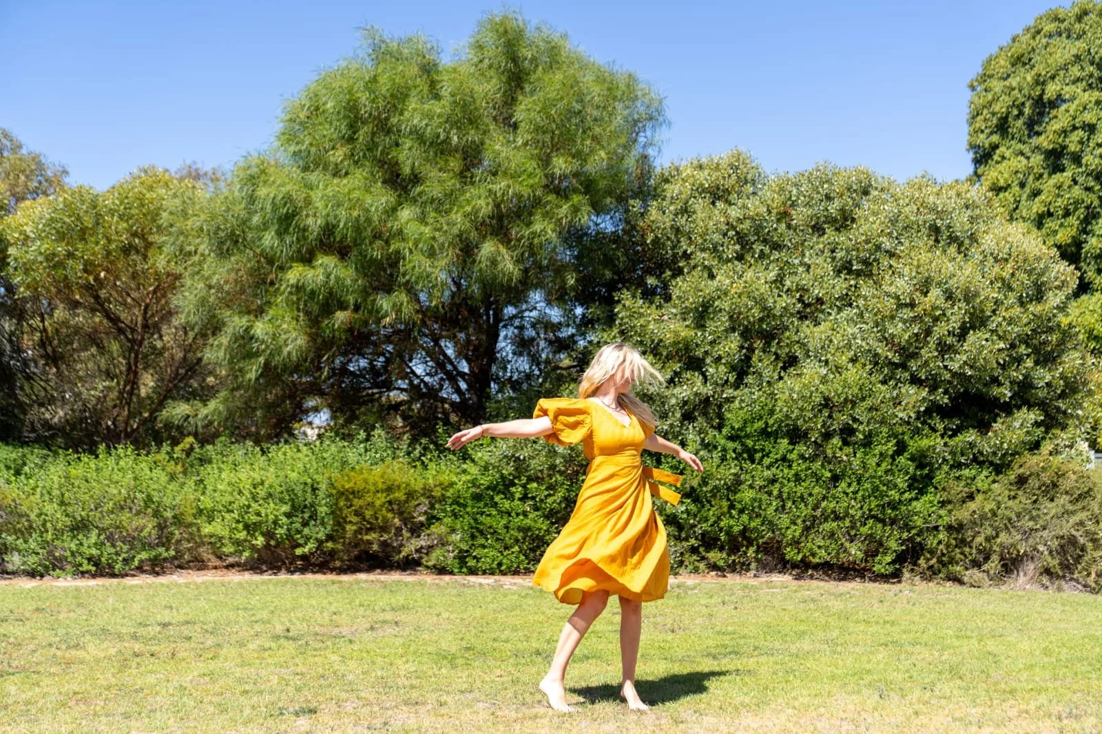 A woman in a yellow dress spinning outdoors on grass with trees and greenery in the background.