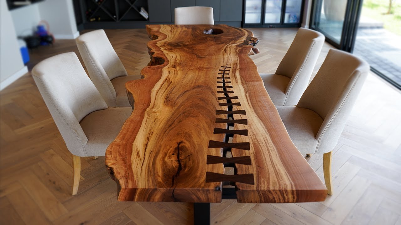 A wooden dining table with a live edge and visible wood grain, featuring a decorative inlay resembling a railroad track, surrounded by six beige upholstered chairs in a modern dining room with large windows.