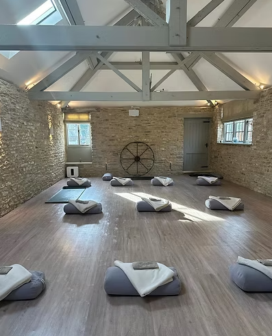 A Barre class studio with wooden floors, exposed brick walls, and a large wheel decoration on the back wall. Ten mats with folded towels are evenly spaced on the floor, arranged in a semi-circle. The room has skylights and windows with natural light.