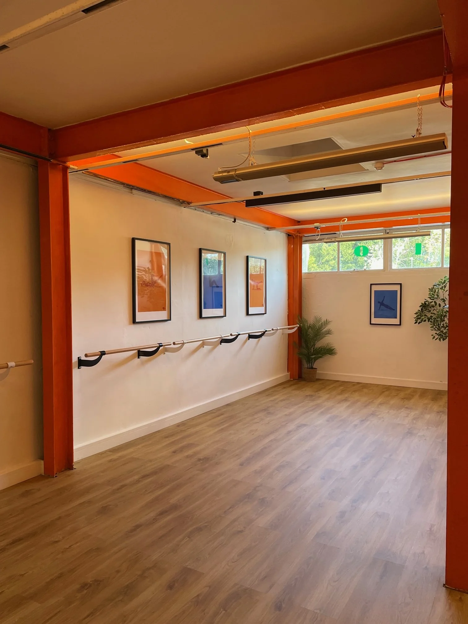 An empty Barre Class studio with wooden flooring, orange and white walls, framed pictures on the wall, a horizontal ballet barre, a window with greenery outside, and ceiling lights.