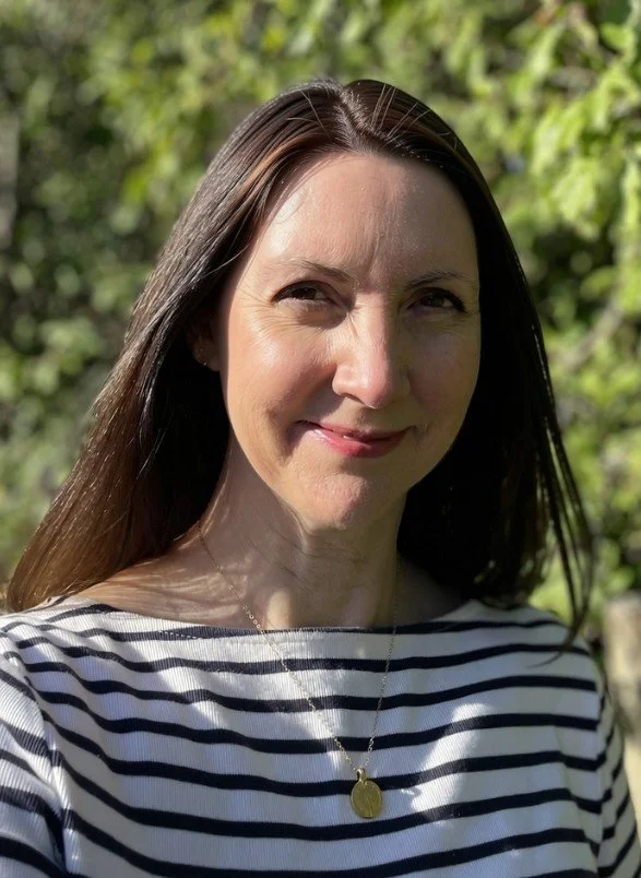 A woman called Claire, who set up WonderBarre Wellness. The woman has long brown hair, is smiling outdoors, wearing a navy and white striped shirt and a gold necklace, with green foliage in the background.