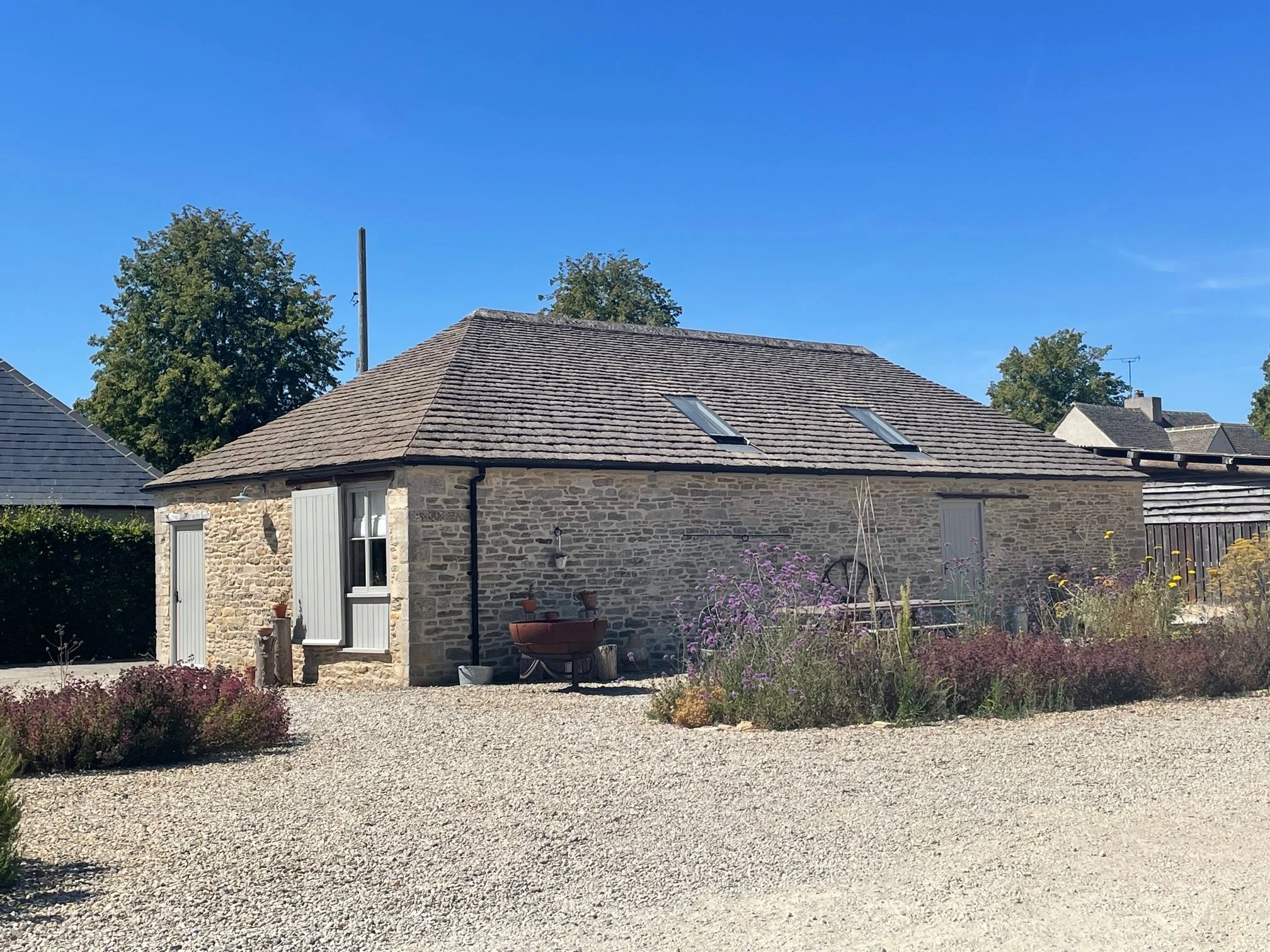A converted stone barn with a sloped wooden roof, two skylights, a window with shutters, and a gravel yard with plants and flowers, under a clear blue sky.