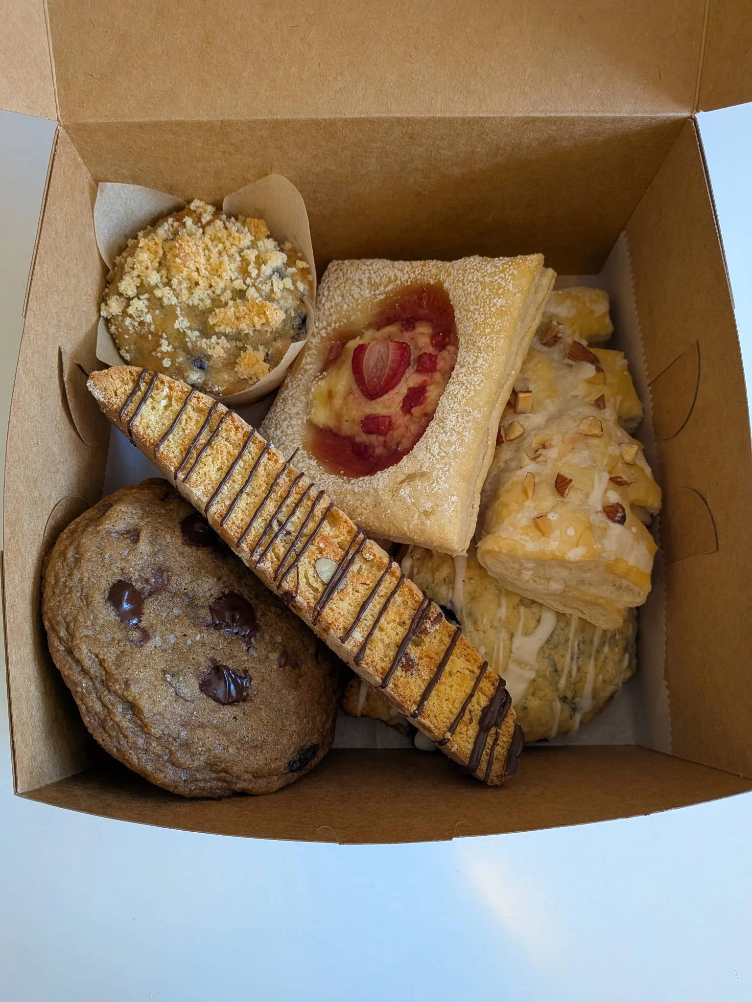 Assorted baked goods in a brown cardboard box, including a cookie with chocolate chips, a crumb-topped muffin, a slice of cherry-filled pie, a slice of yellow cake with nuts on top, and a biscotti with chocolate drizzle.