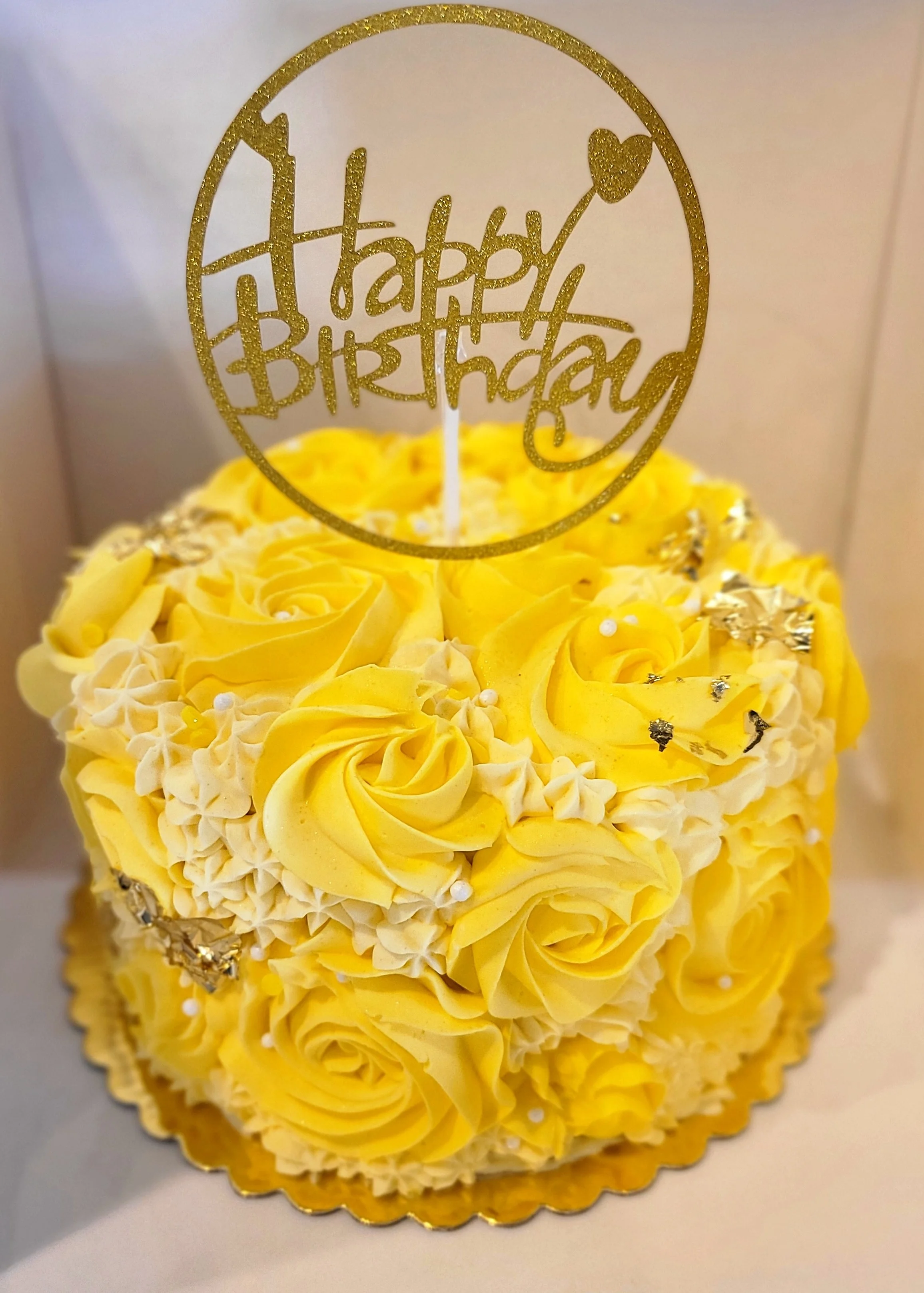 A yellow and white birthday cake decorated with yellow icing roses, silver beads, and Gold Elements. A gold glittery cake topper reads 'Happy Birthday' inside a circle with a heart-shaped balloon.