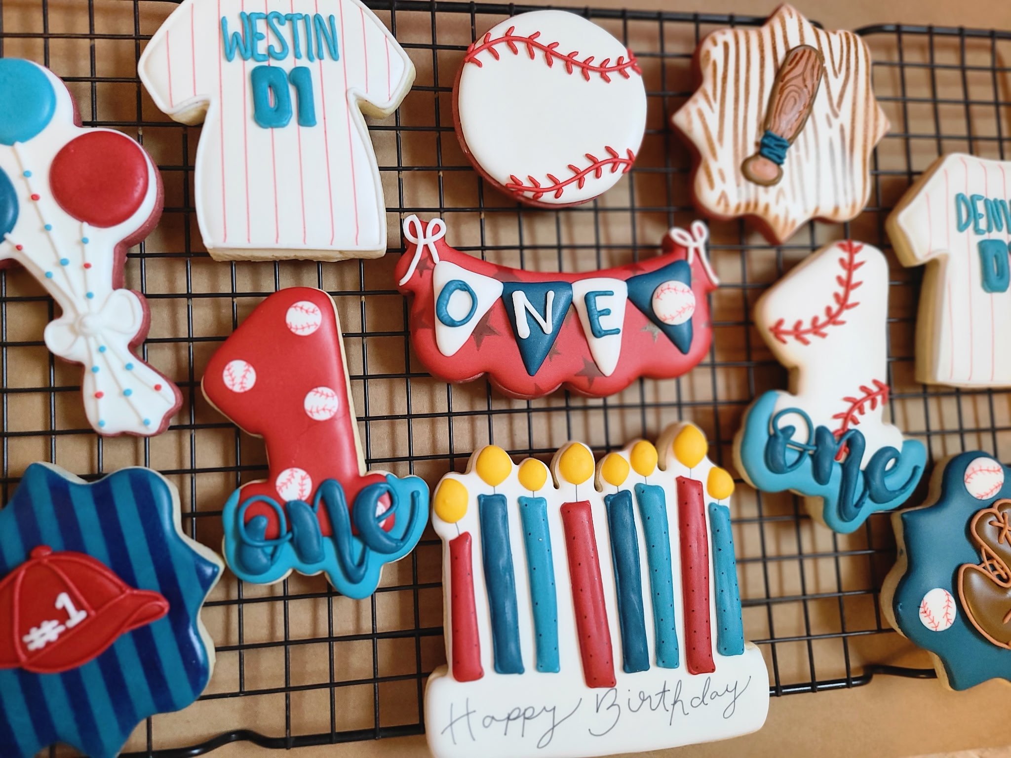 Decorative cookies on a wire rack, themed for a baseball or sports birthday celebration, including cookies shaped like a baseball, a jersey with 'WESTIN 01', a star with a baseball bat, and a banner reading 'ONE'.