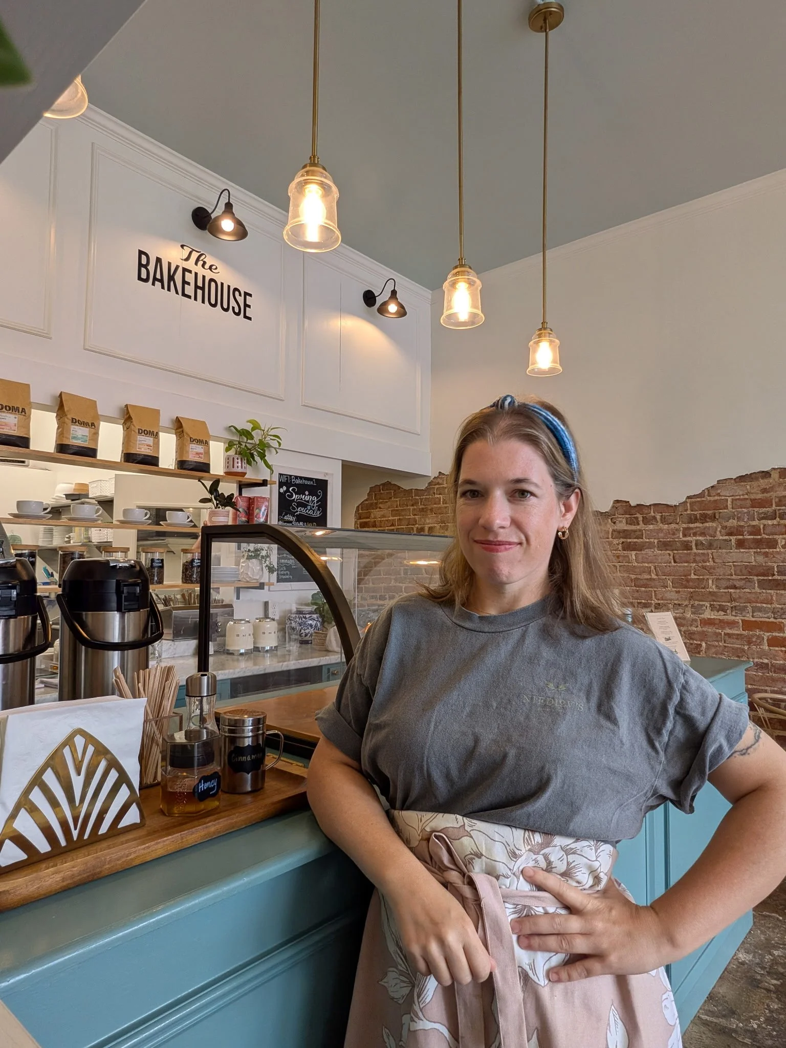 A woman standing at a cafe counter in front of a brick wall and a sign that says "The Bakehouse." She is wearing a gray t-shirt with a logo on it and an apron, with her left hand on her hip and smiling.