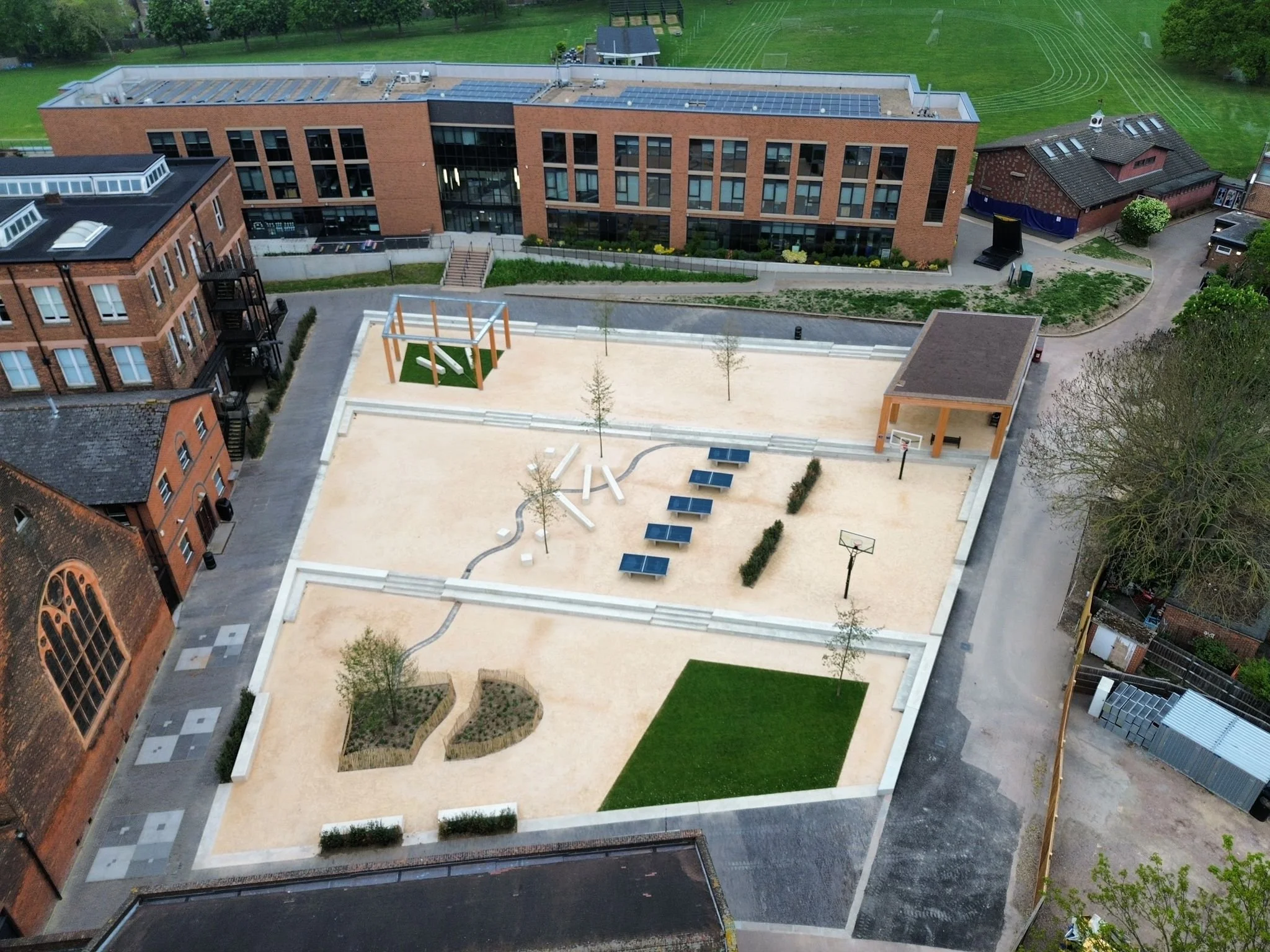 Aerial view of a modern brick building with large glass windows, surrounded by a playground, a small basketball court, a grassy field, and trees.