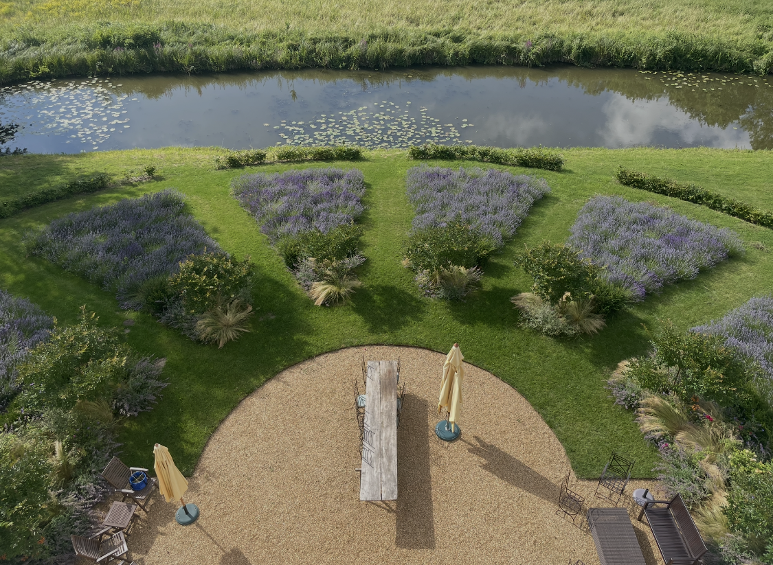 A backyard garden with a pond in the background and a landscaped area with purple flowers and trees in the foreground. There is a gravel patio with outdoor furniture, umbrellas, and a long wooden table.